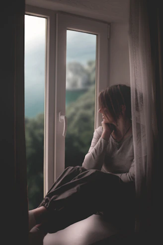 A woman sits by a window, gazing out.