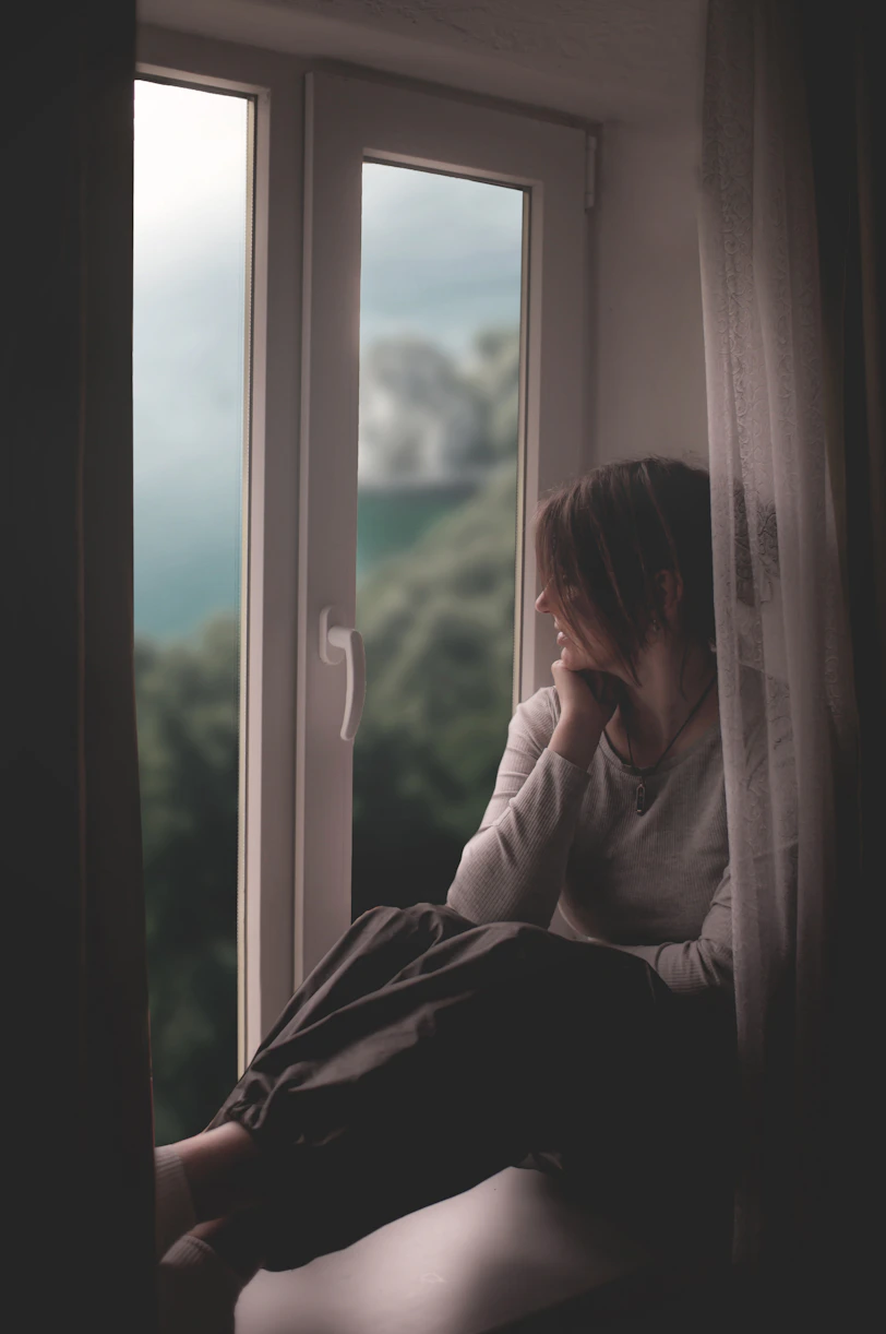 A woman sits by a window, gazing out.