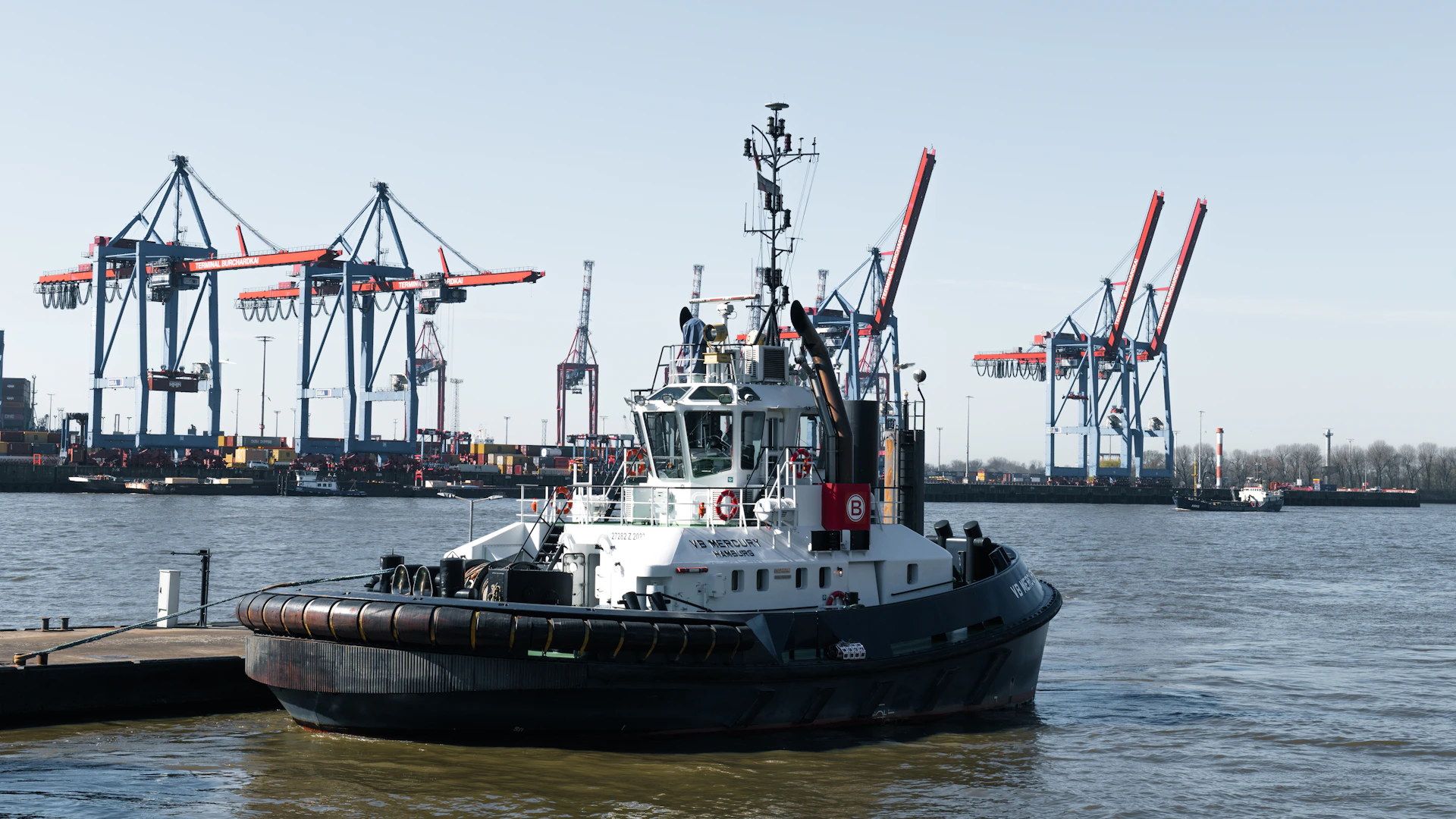 A tugboat sails by dockside cranes