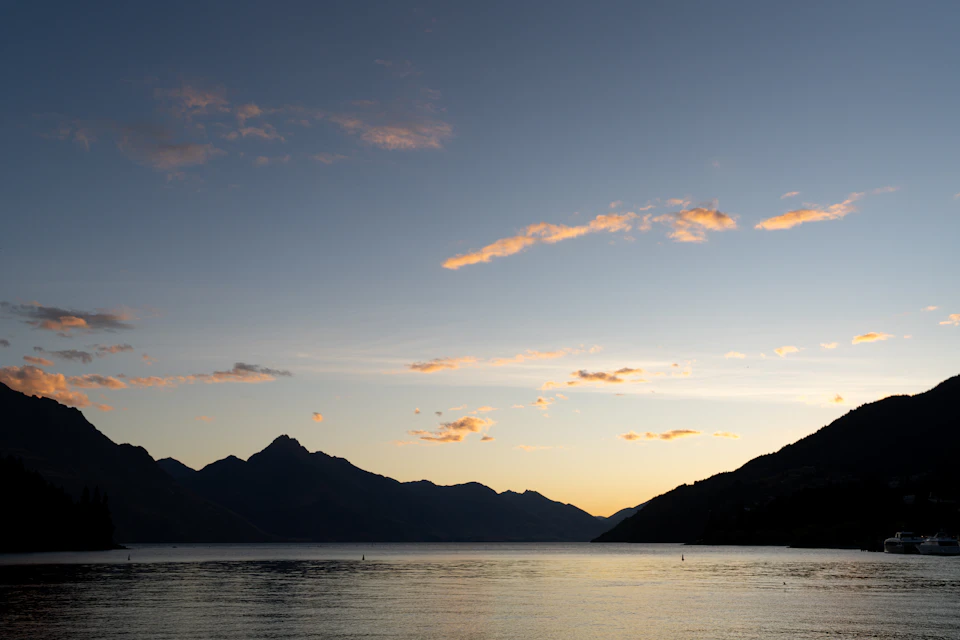 The image depicts a serene sunset over a mountain range, with the sun setting behind the mountains and casting a warm glow across the sky. The mountains are silhouetted against the vibrant orange hues of the setting sun, creating a breathtaking backdrop for the scene. The water below mirrors the colors of the sky, adding to the overall beauty of the landscape.