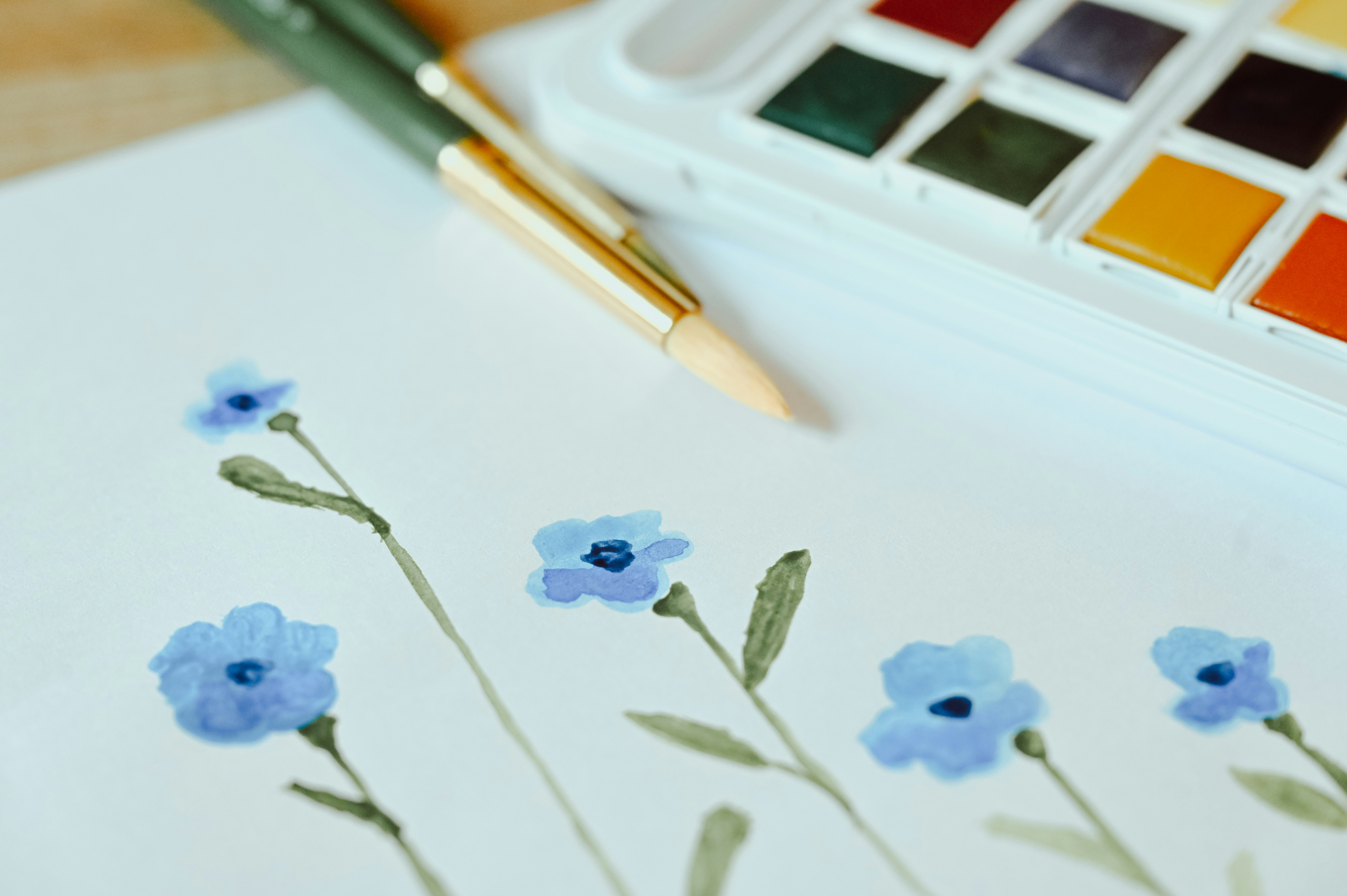 Small blue watercolour flowers near 2 paintbrushes and a paintbox.