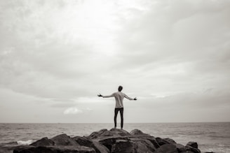 Man stands on rocks, arms outstretched towards the sea.
