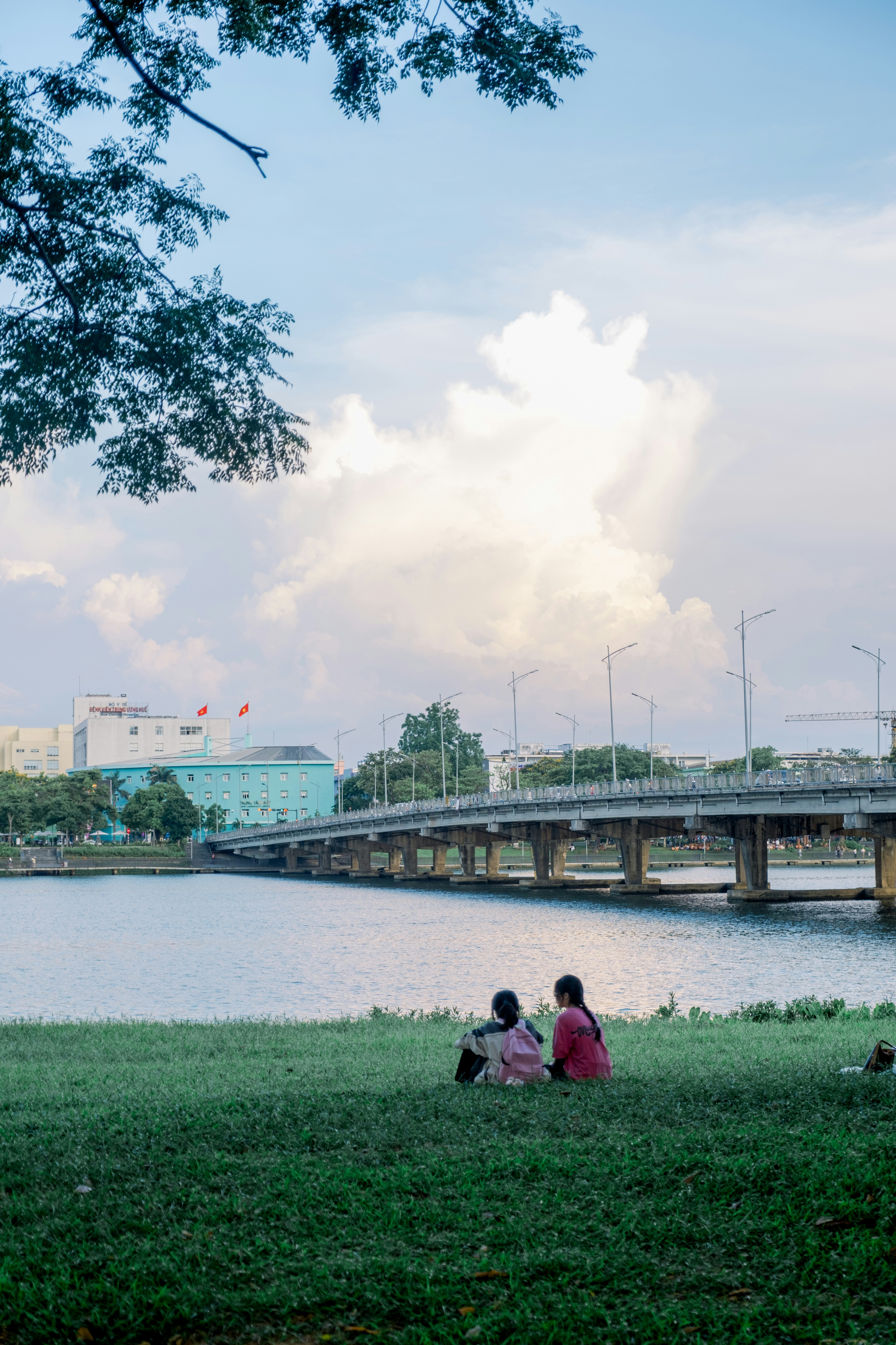 Two people sit by the water, watching the bridge.