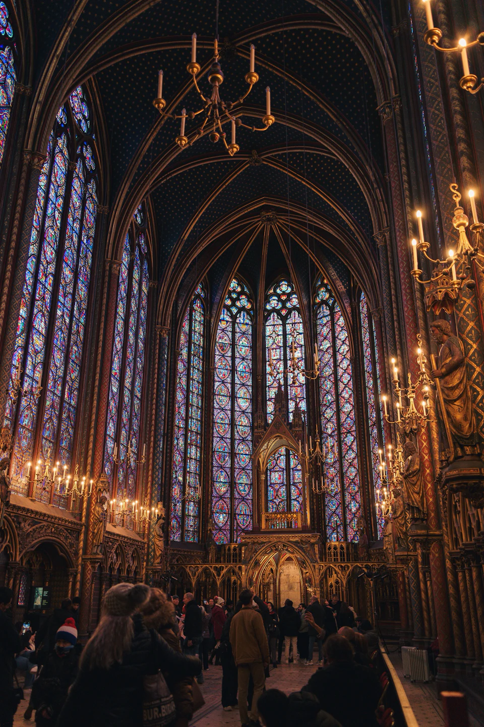 The image depicts a large, ornate church with a high ceiling and numerous stained glass windows that cast colorful light throughout the space. The walls are adorned with gold accents, adding to the grandeur of the scene. A chandelier hangs from the center of the room, illuminating the area below. Several people can be seen walking around the church, some carrying bags or wearing backpacks.