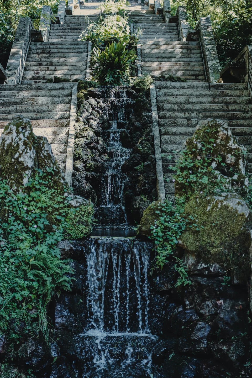The image depicts a serene waterfall cascading down a series of stone steps, surrounded by lush greenery and rocks. The water is crystal clear, reflecting the surrounding environment. The perspective of the photo is from below, looking up at the waterfall, creating a sense of awe and wonder. The colors in the image are predominantly green and brown, with the clear blue sky providing a striking contrast to the earthy tones of the stone steps and vegetation.

The image does not contain any discernible text or countable objects, making it an interesting visual experience for viewers.