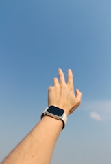 Hand with smartwatch reaches towards the blue sky.