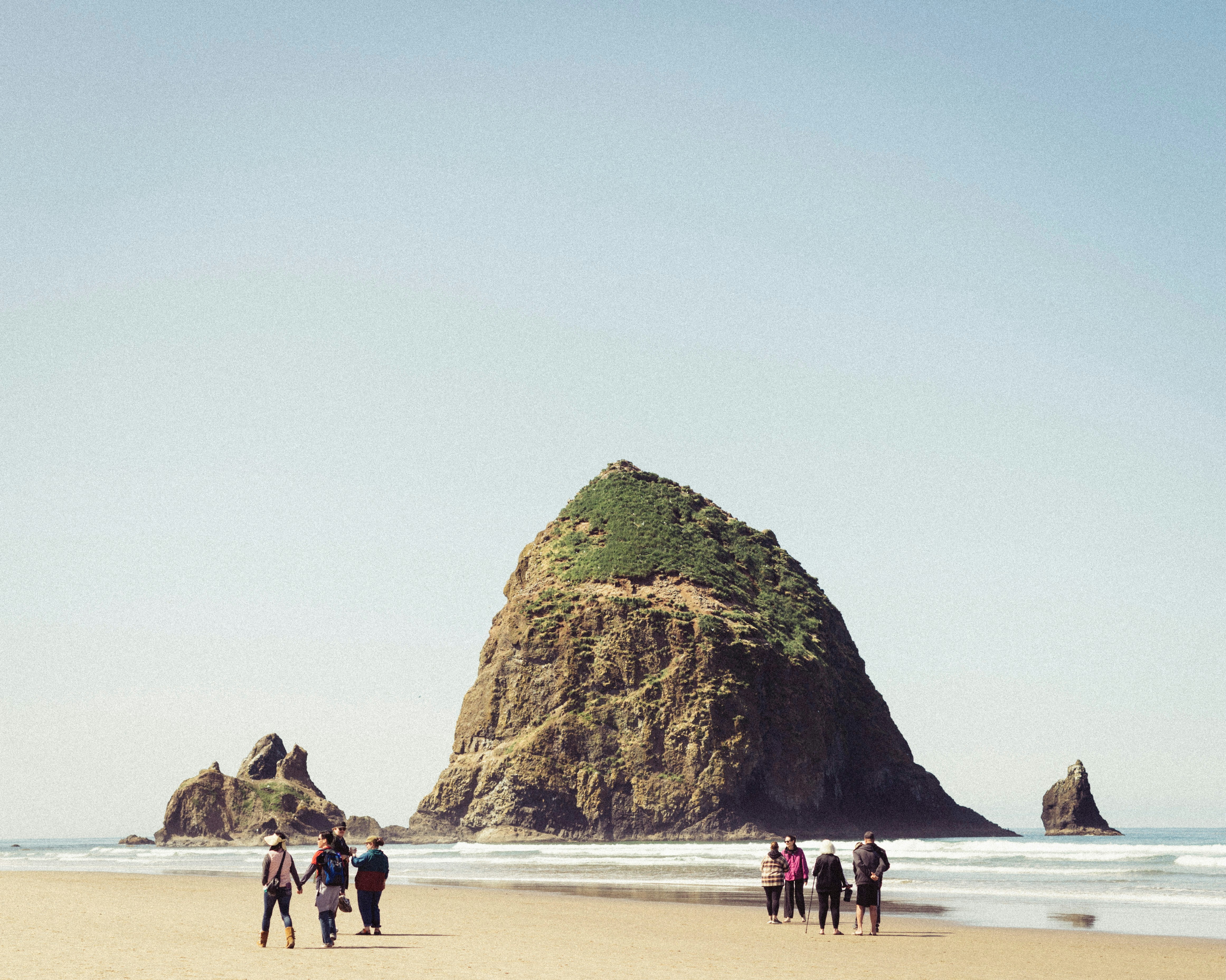 People stroll on a sandy beach near a large rock.