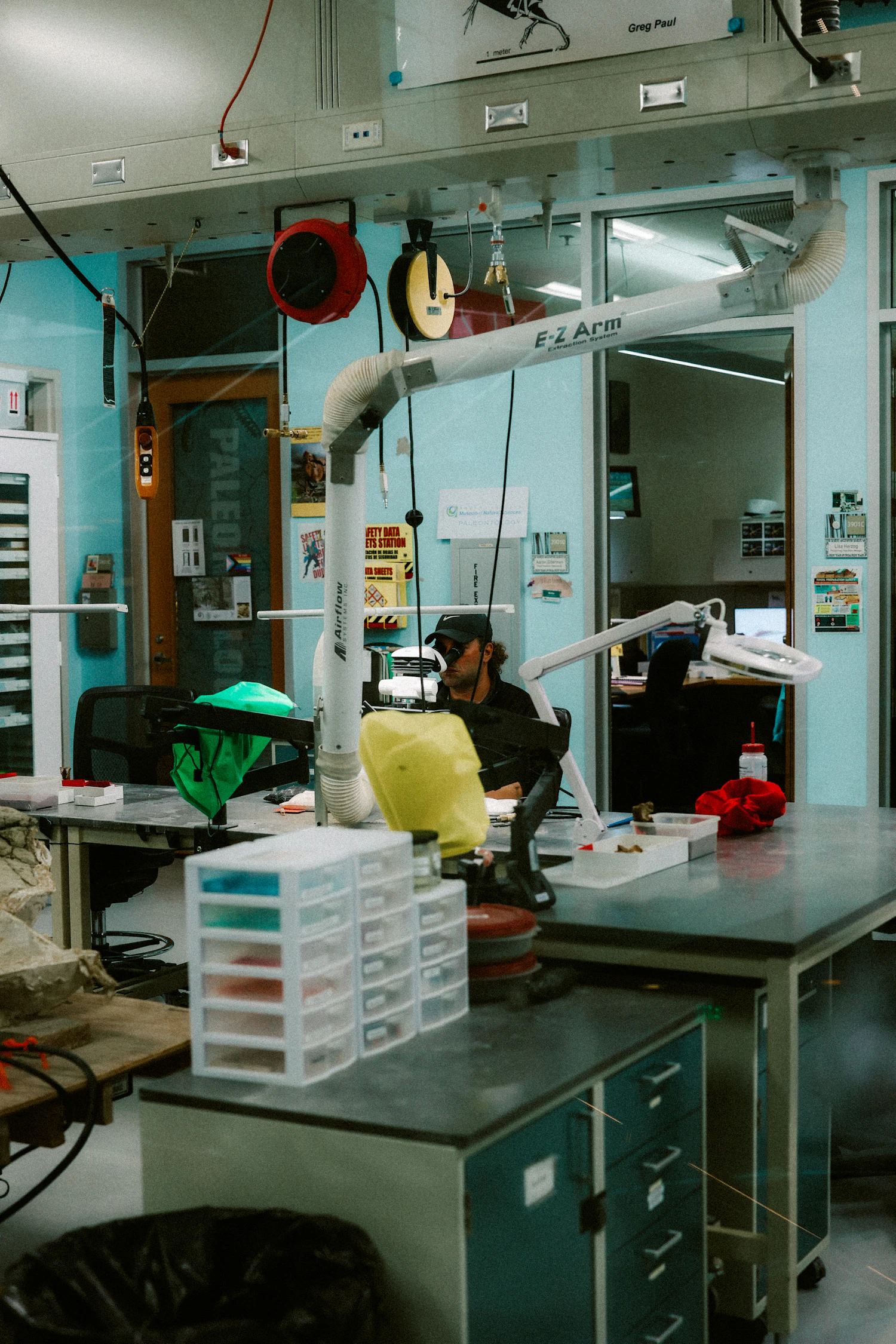 A cluttered repair bench under a magnifier lamp, drawers of parts and a seated technician in the background