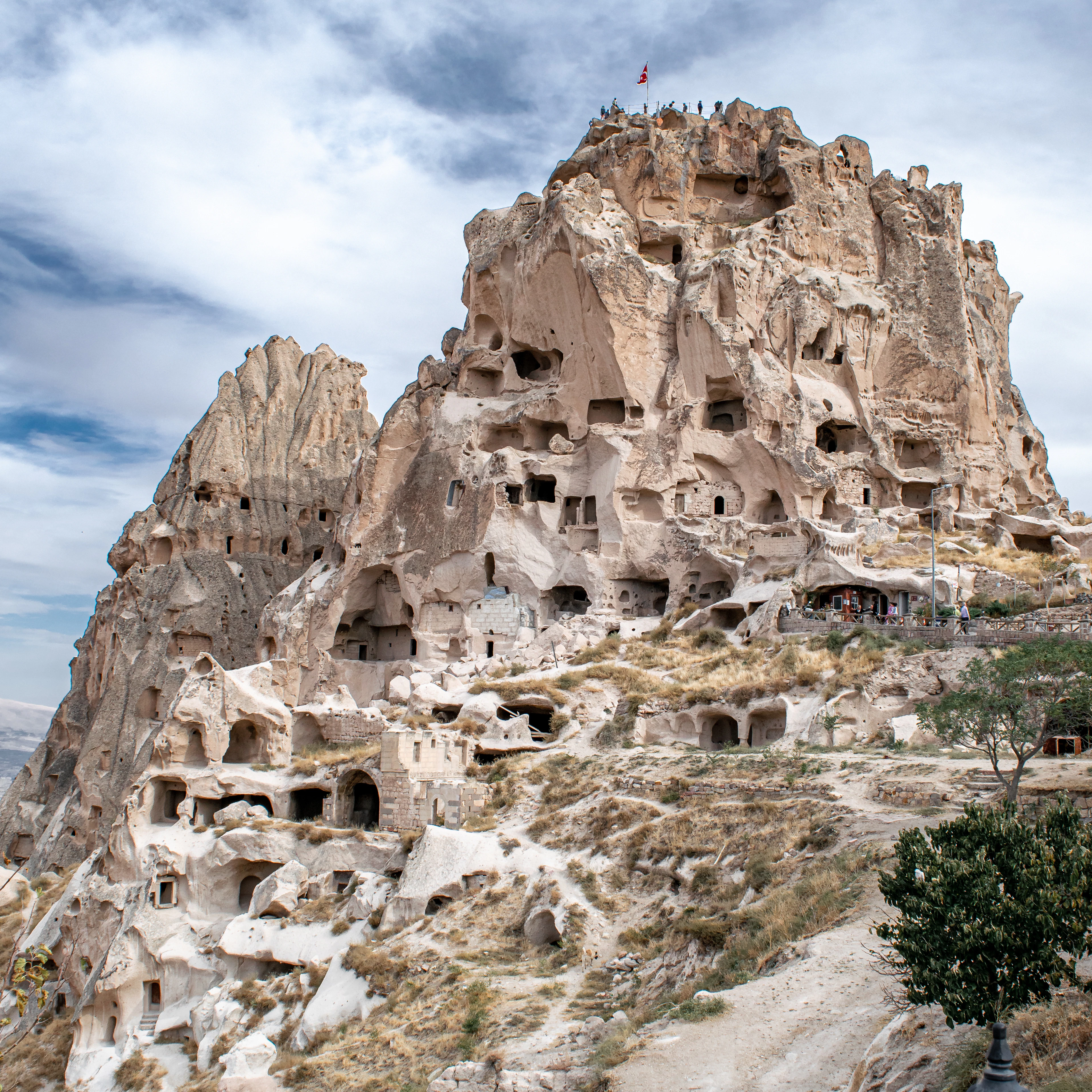 Uchisar Castle in Cappadocia, a towering pinnacle of pale tuff honeycombed with hundreds of hand-cut windows and chambers, a Turkish flag flying from the summit where visitors stand, under a sky of dramatic broken cloud
