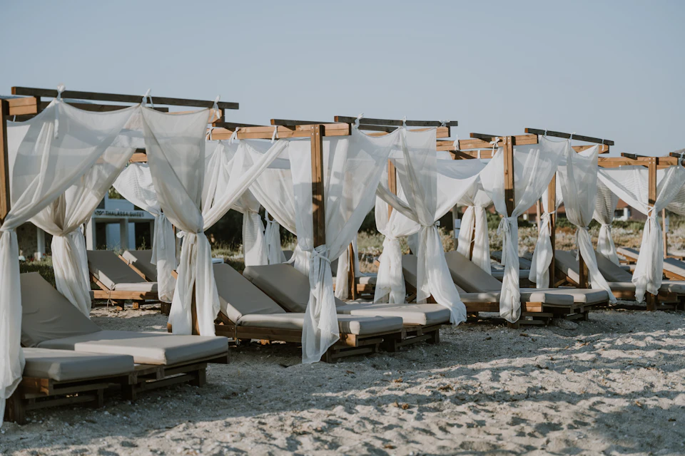 Rows of lounge chairs and beds on the sand with white curtains swaying in the