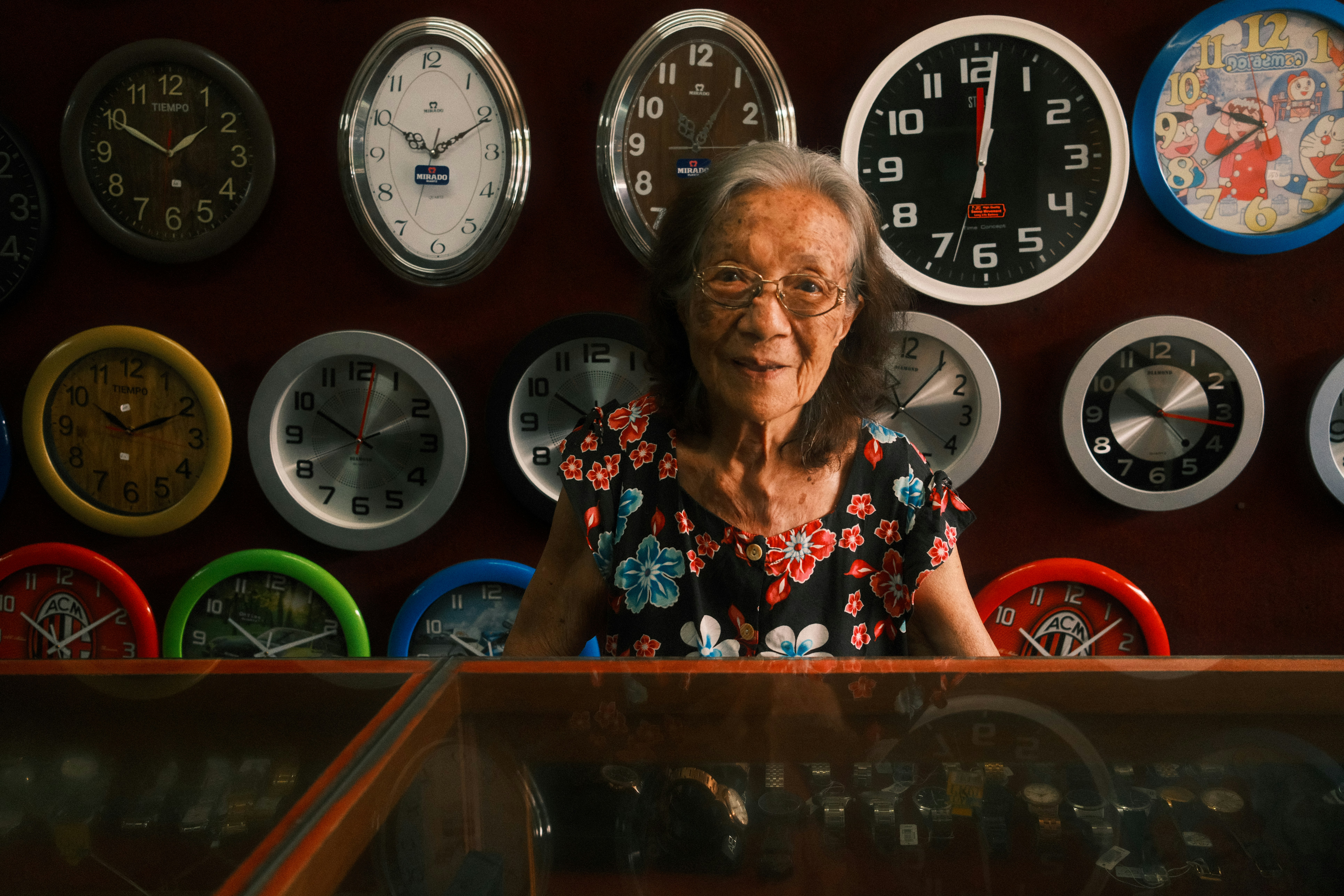 Elderly woman smiles in front of many clocks.