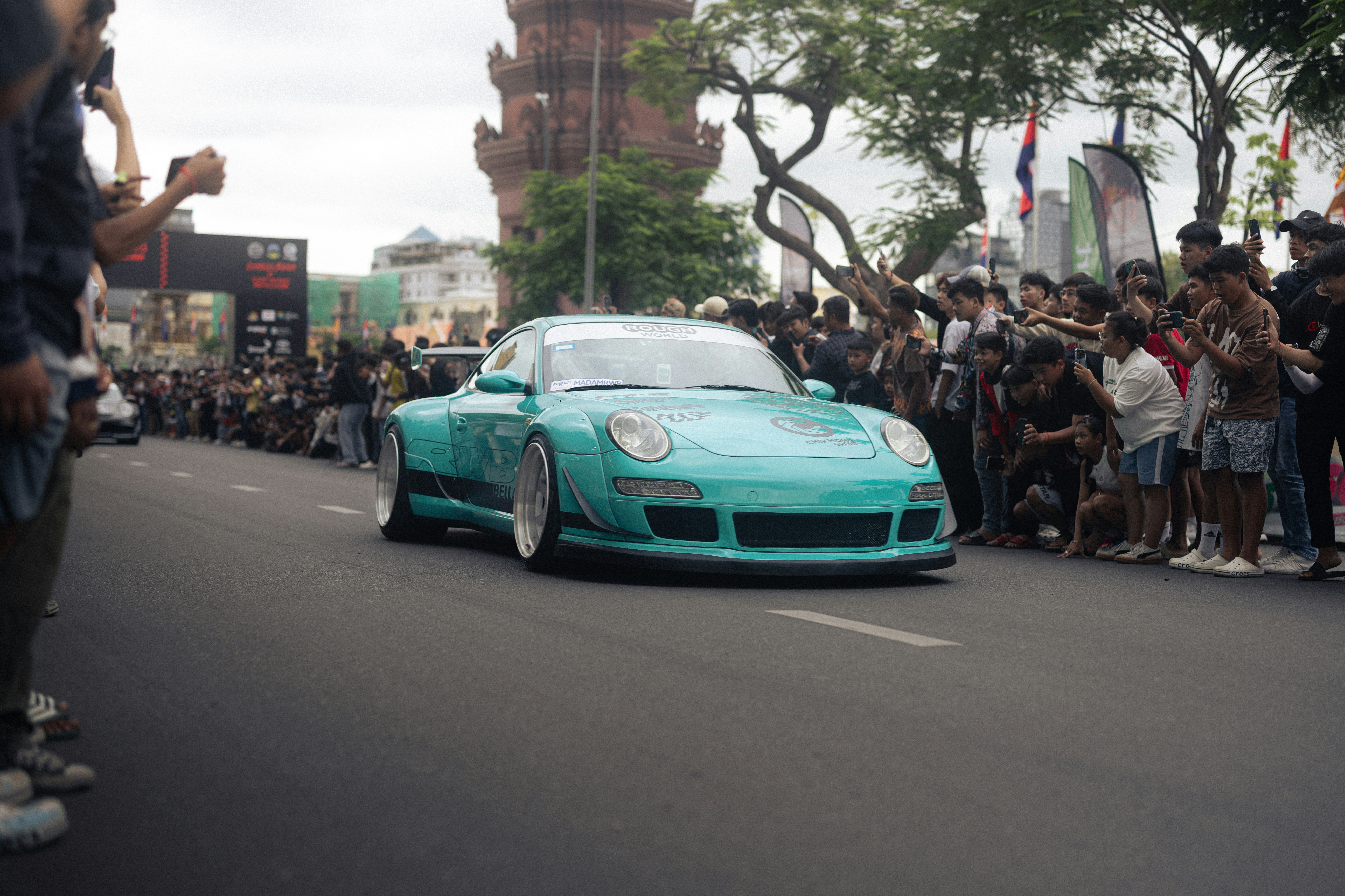 A turquoise sports car drives past many people.