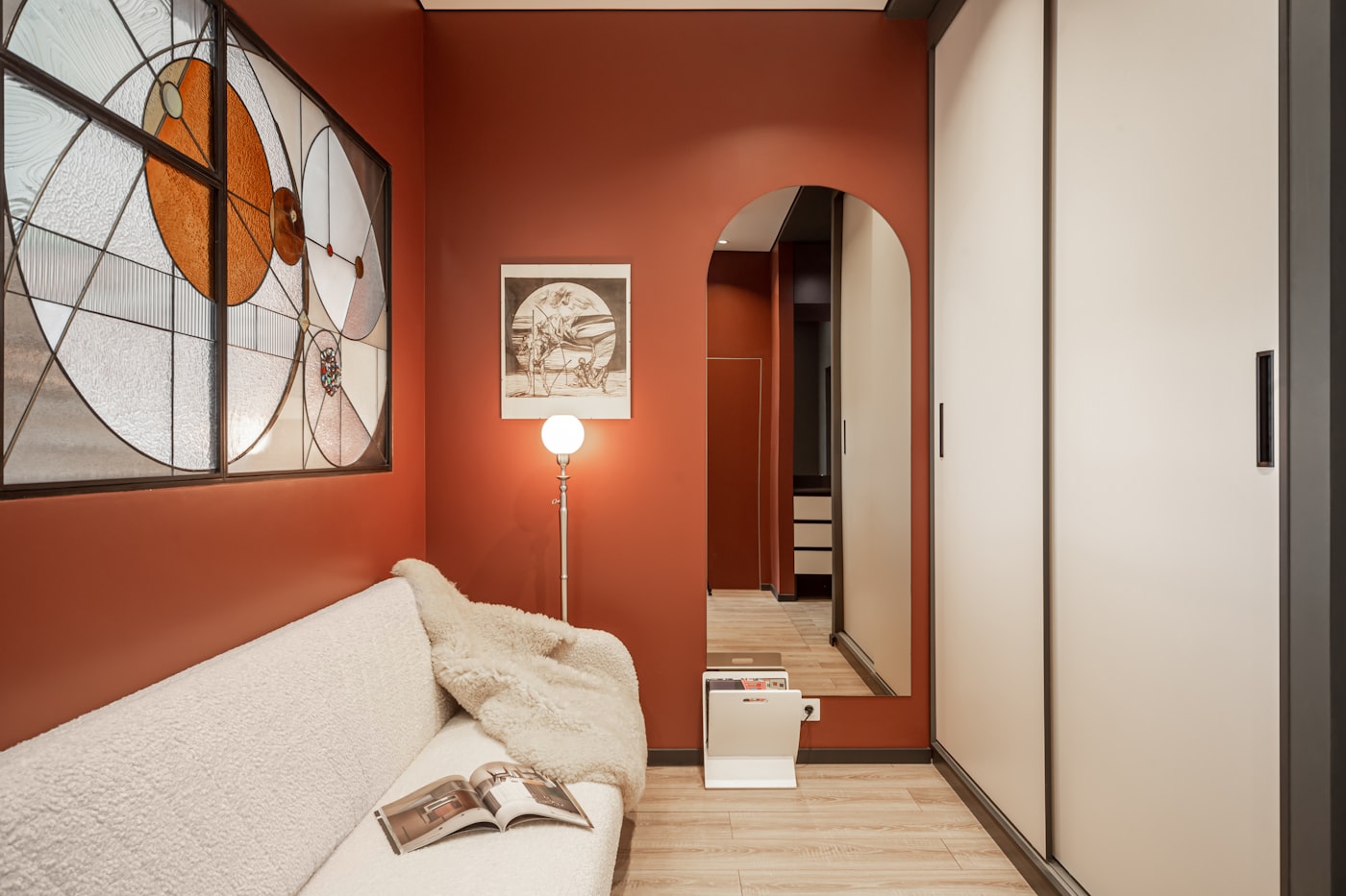 Cozy small apartment room with deep terracotta red walls, arched doorway, cream bouclé sofa, stained glass window, and warm floor lamp creating an intimate earthy atmosphere