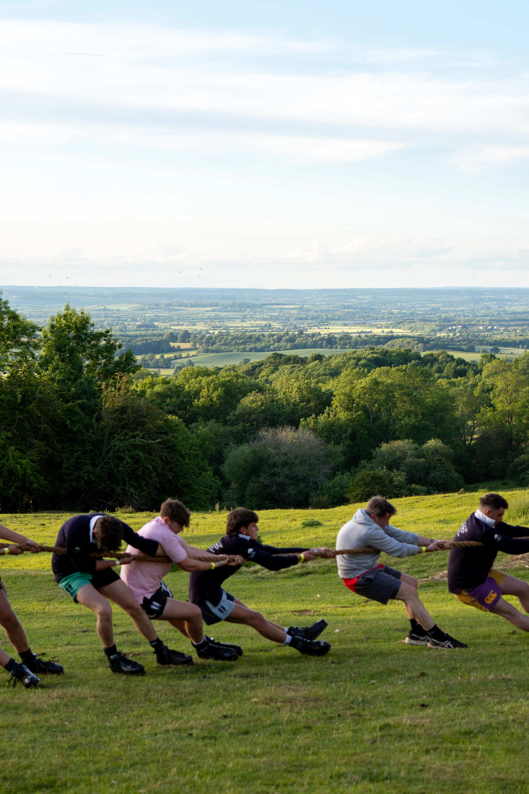 Five pullers leaning low on a rope on a grassy hillside under a wide evening sky