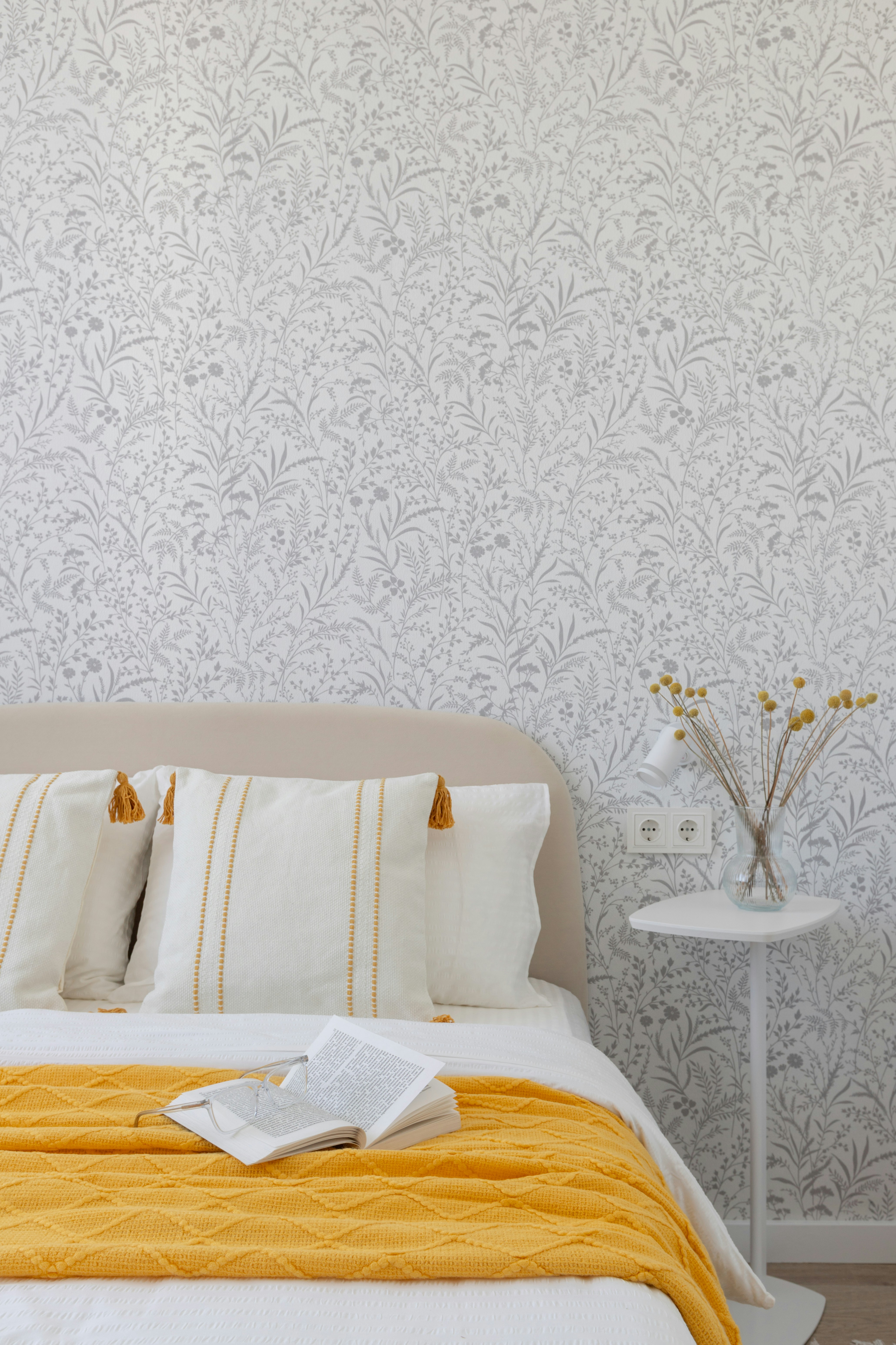 A cozy bedroom features book and floral wallpaper.