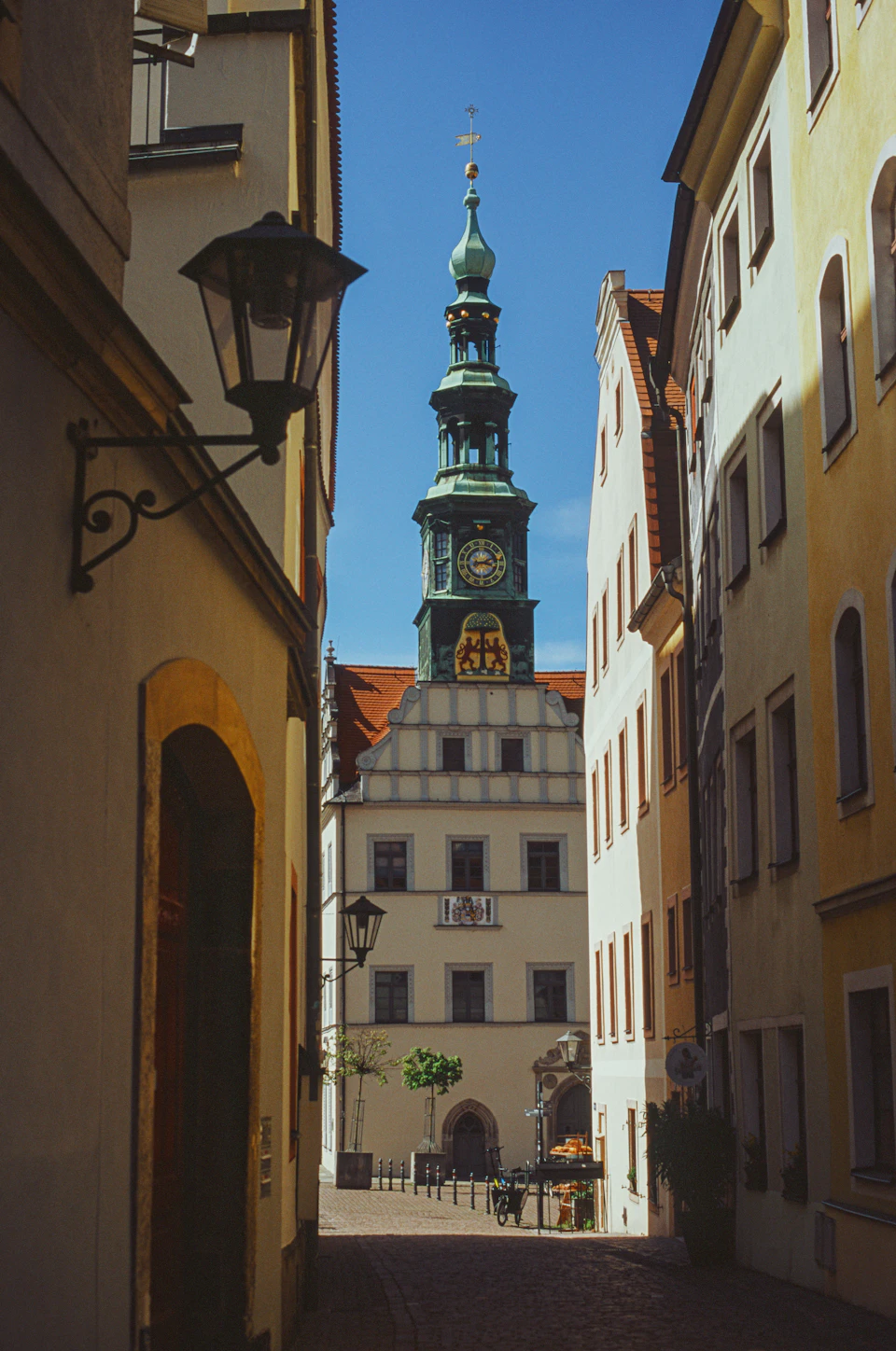 The image depicts a narrow cobblestone street flanked by old buildings with yellow and white facades, including one building on the left that has a green clock tower reaching towards the clear blue sky. The street is lined with trees, adding to the picturesque scene. On the right side of the street, there are two benches where people can sit and enjoy the view.