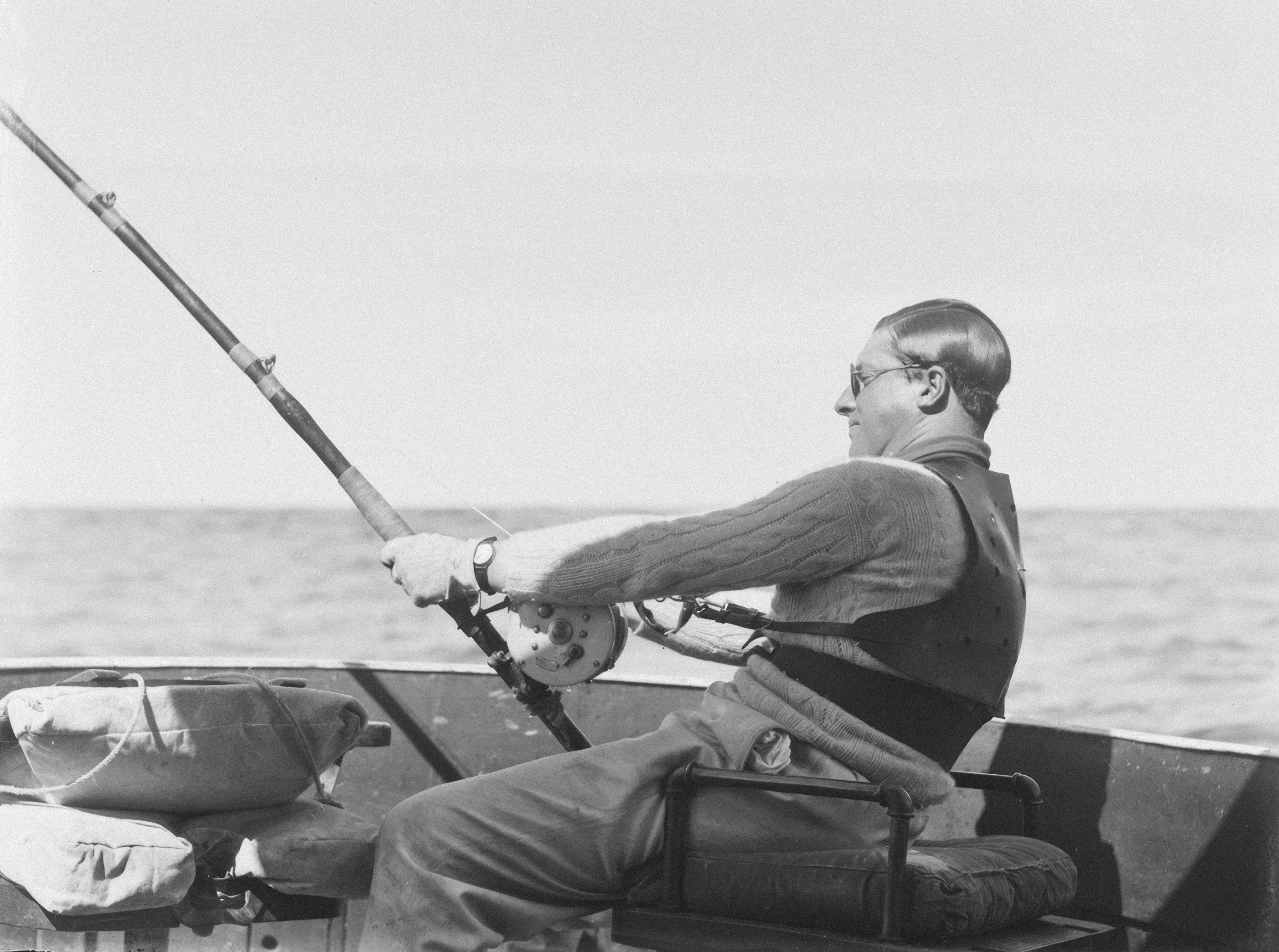 Black-and-white photo of an angler seated at the gunwale of a boat, leaning back in a padded back harness with its straps clipped to a big-game reel and a bent rod