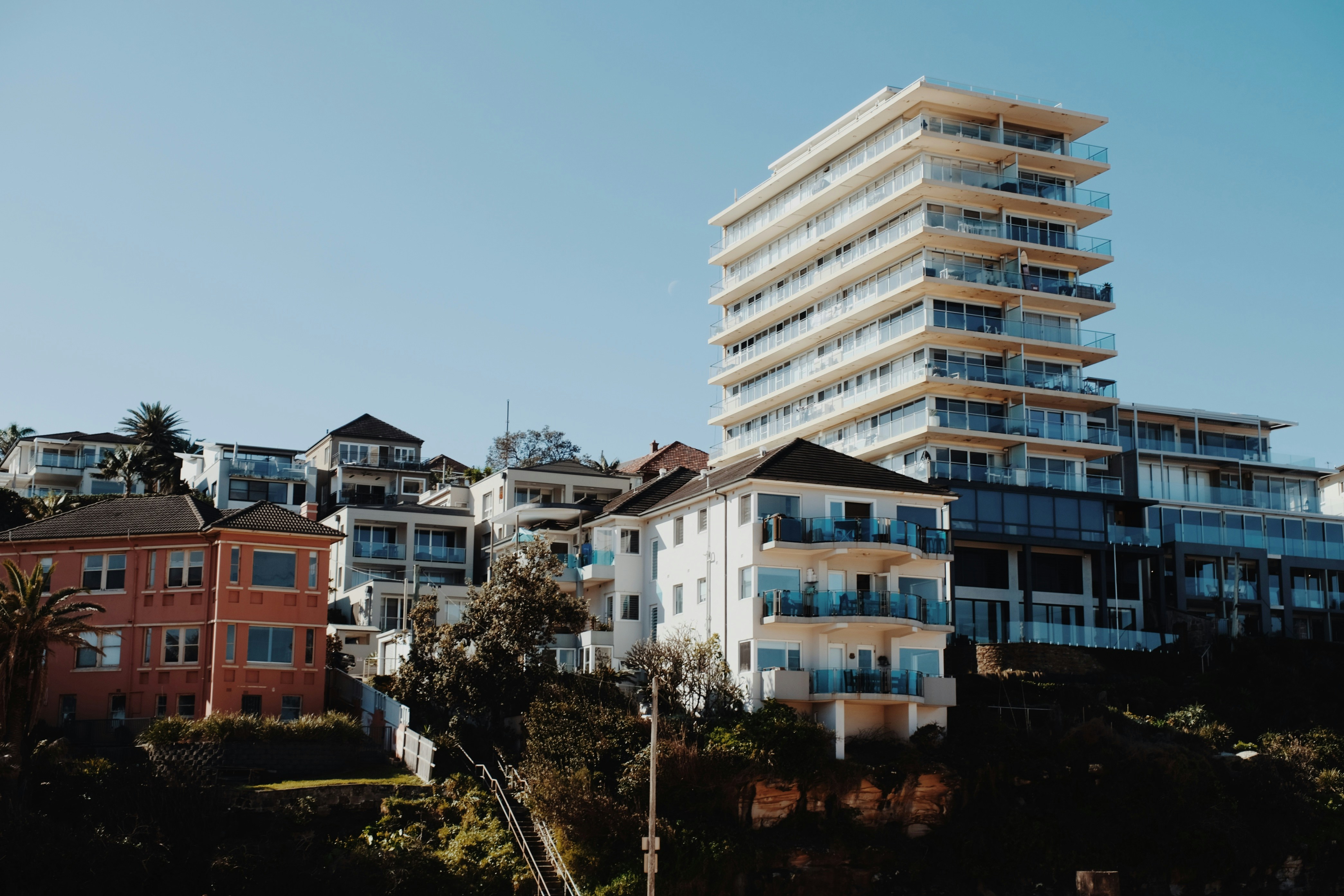 Various houses and a large modern apartment building on a sunny hillside against a clear blue sky, featuring mixed architectural styles.