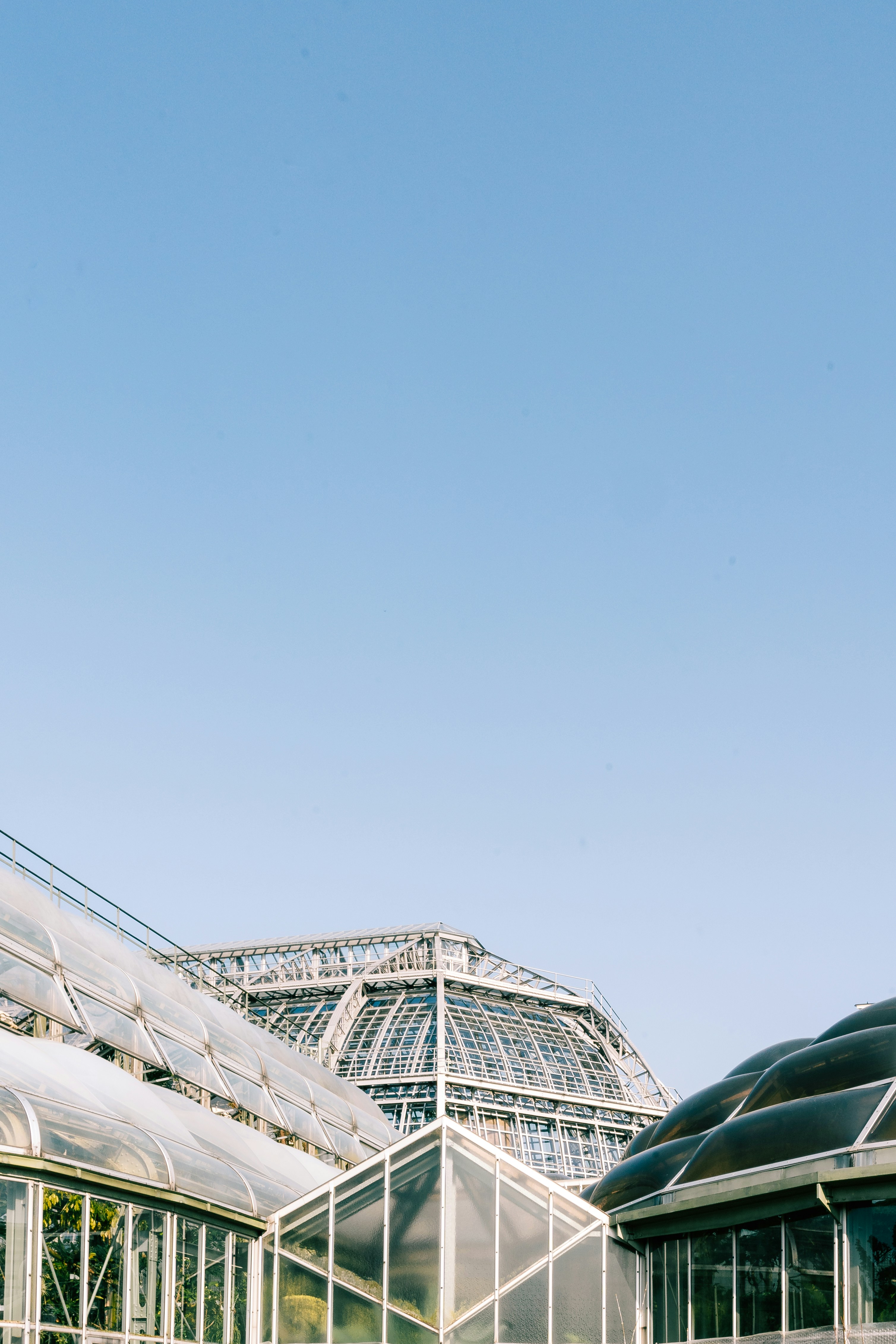 crisp, sunny day casts soft light on this striking cluster of greenhouses, each with its own unique geometry—domes, angular peaks, and steel-framed glass. It almost feels like a sci-fi botanical garden or a research station from the future. The clean lines and transparent structures create a beautiful harmony with the bright blue sky above.