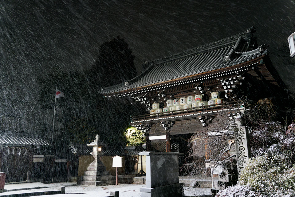 A Kyoto temple stands in light snowfall at night, illuminated against the dark winter sky with snow accumulating on the tile roof and stone lanterns