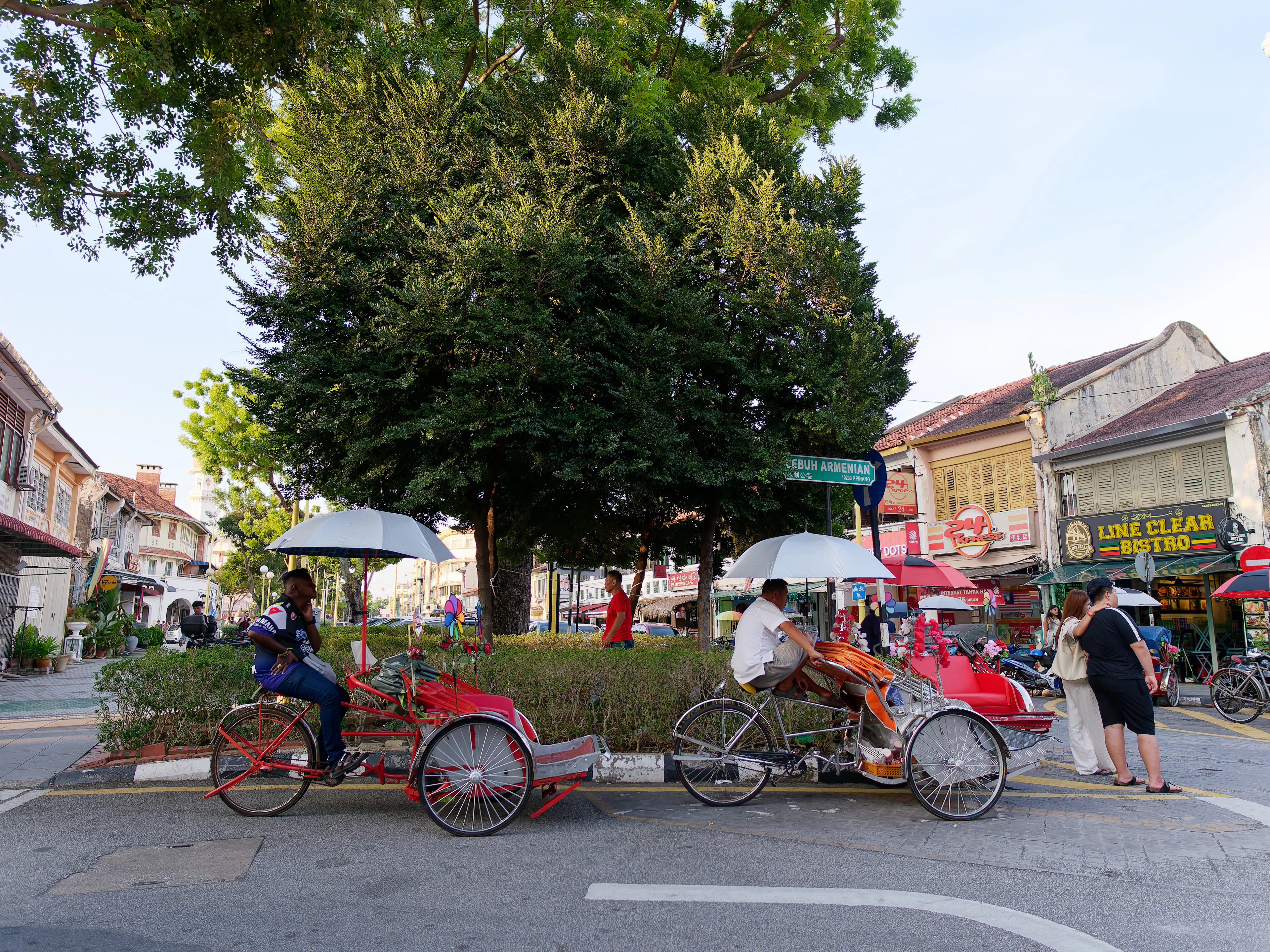 Canopied trishaws parked beside a leafy square in George Town, Penang, with heritage shophouses and shop signage lining the street behind