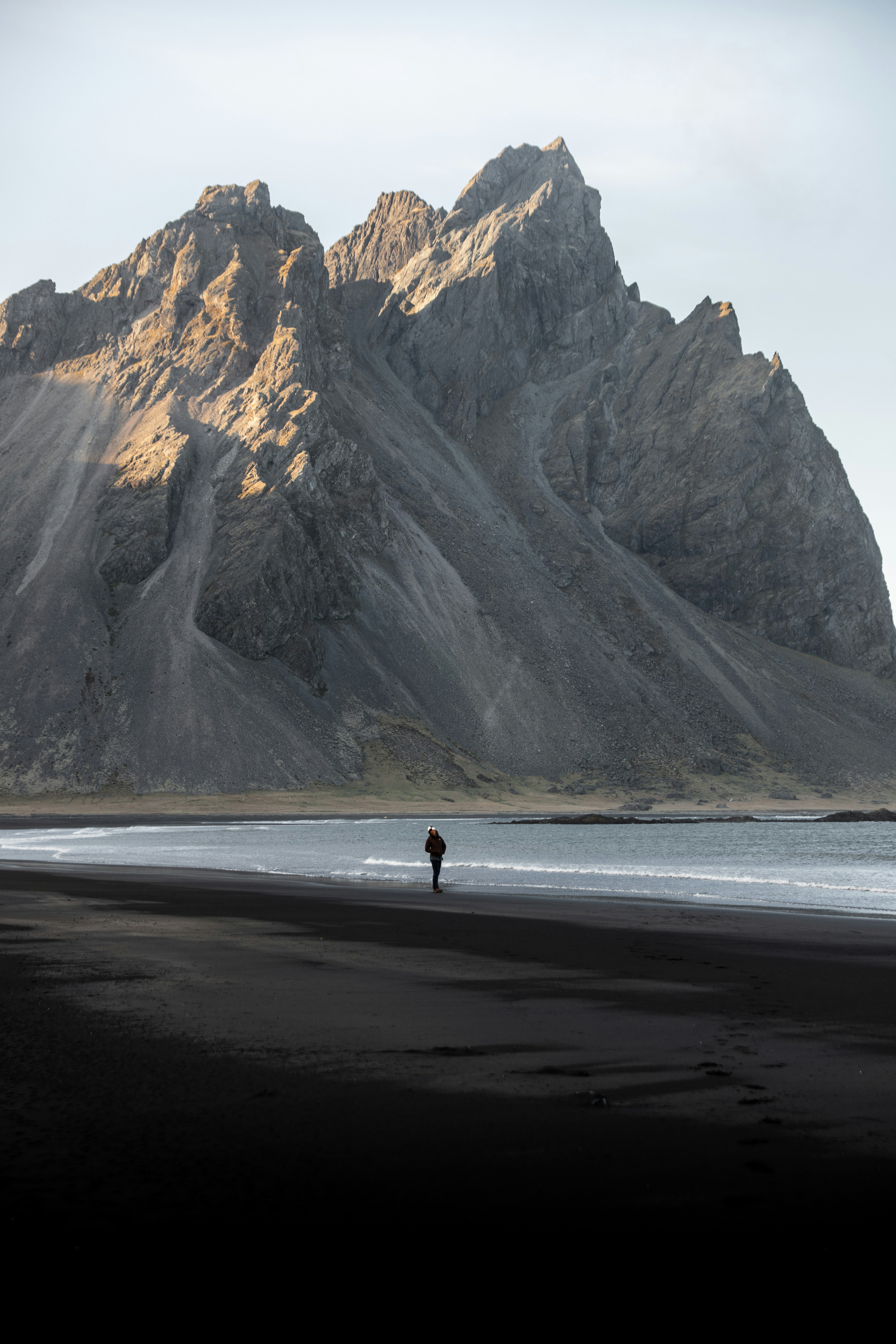 Person stands on a black sand beach near a mountain.
