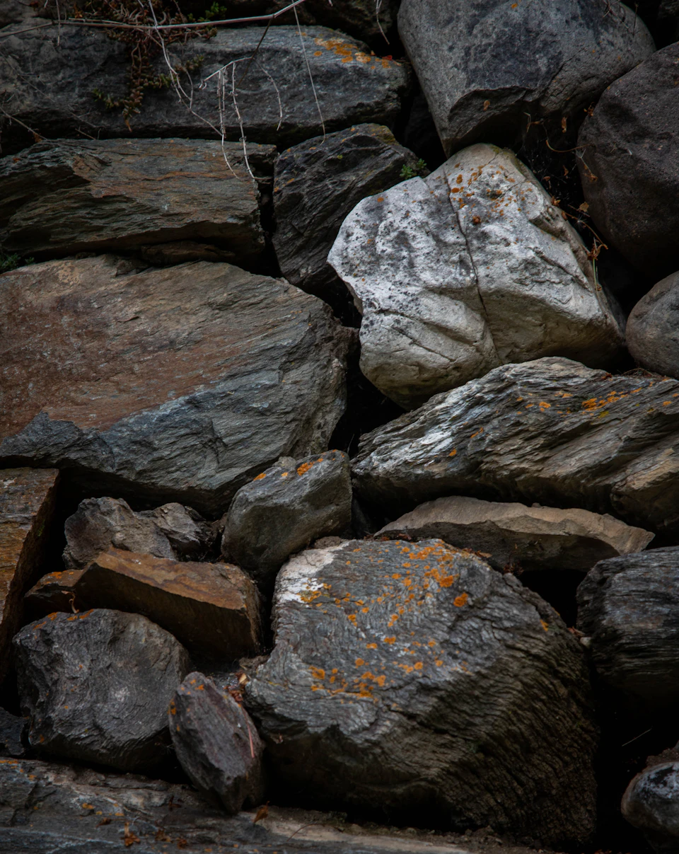 The image shows a close-up view of a rocky cliff face, with various types of rocks and boulders scattered across it. The largest rock is positioned on the right side of the frame, while smaller rocks are found in the center and left side of the image. Some of these rocks have patches of lichen growing on them, adding a touch of green to the otherwise gray and brown landscape.