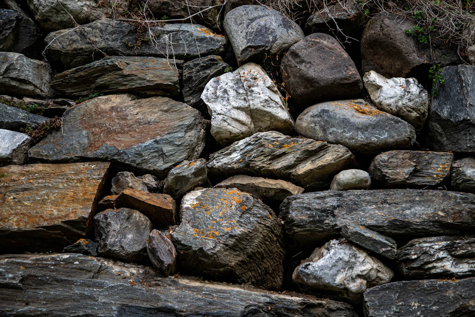 The image shows a close-up view of a rock wall composed of various types of rocks and boulders, including gray, brown, white, and black stones arranged in an uneven pattern. The largest boulder is positioned on the right side of the image, while smaller rocks are scattered throughout the scene. The background appears to be a blurred landscape or a wall, providing context for the rock formation.