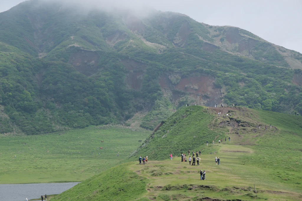Hikers crossing green volcanic grassland below a misty crater ridge at Mount Aso