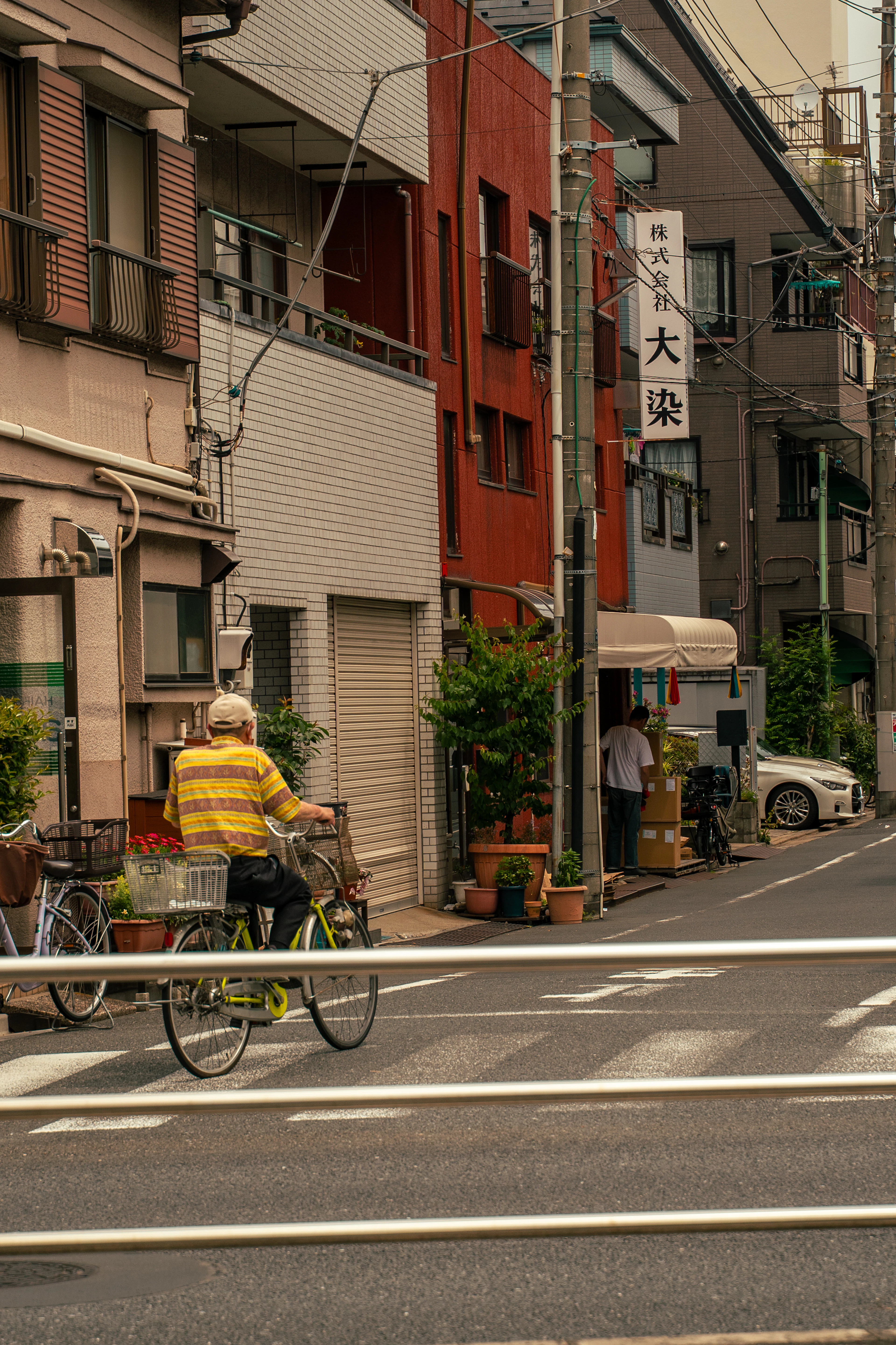 A quiet residential backstreet in Tokyo — a cyclist in a striped shirt, potted plants along the kerb and a vertical shop sign overhead