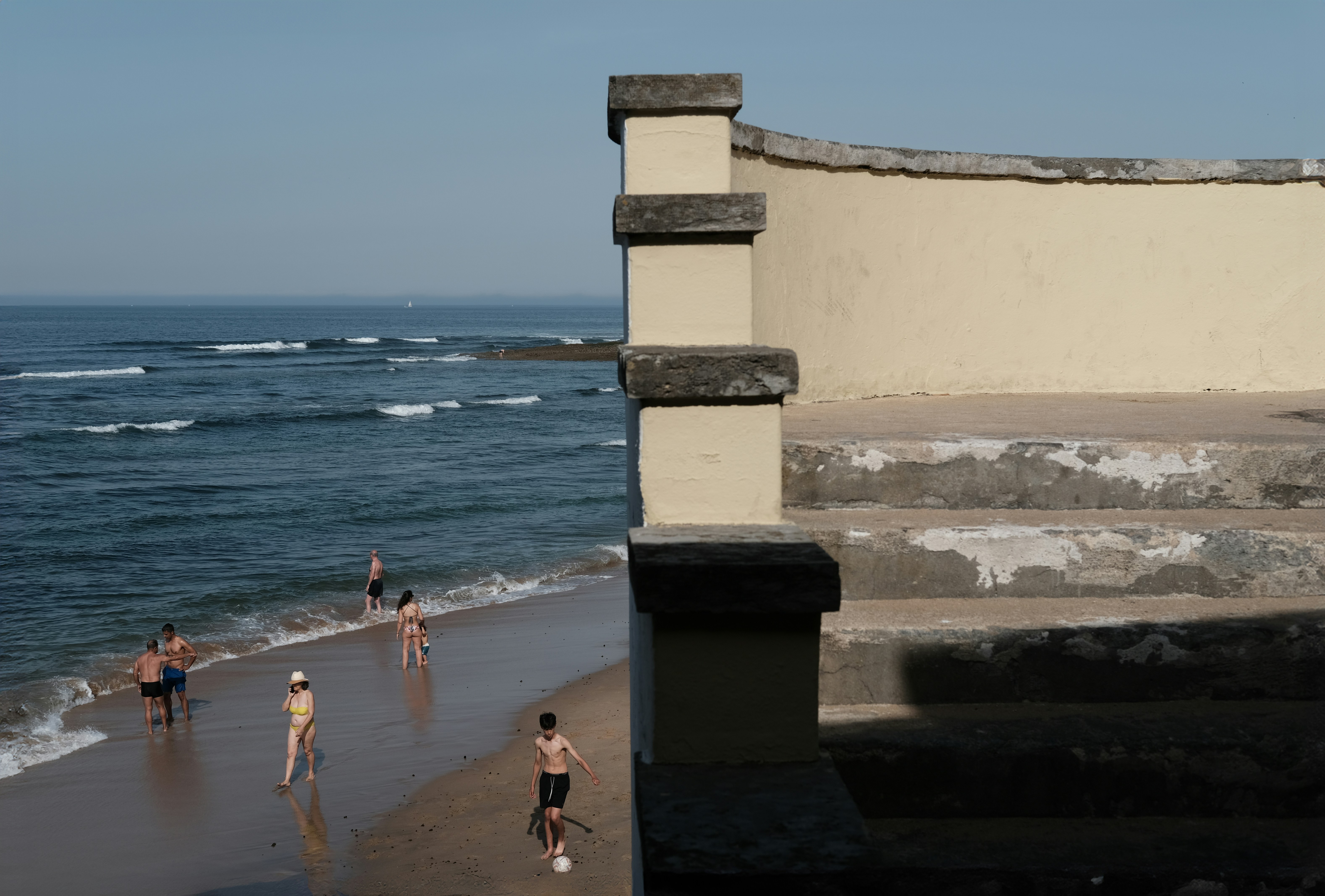 A wall cuts the scene clean in half. On one side, the sea is alive - waves rolling in, light moving across the water, everything shifting and full of motion. On the other, it’s dry and still. A set of old concrete steps, cracked and faded, leading up to a quiet platform. No one’s there. Nothing moves.