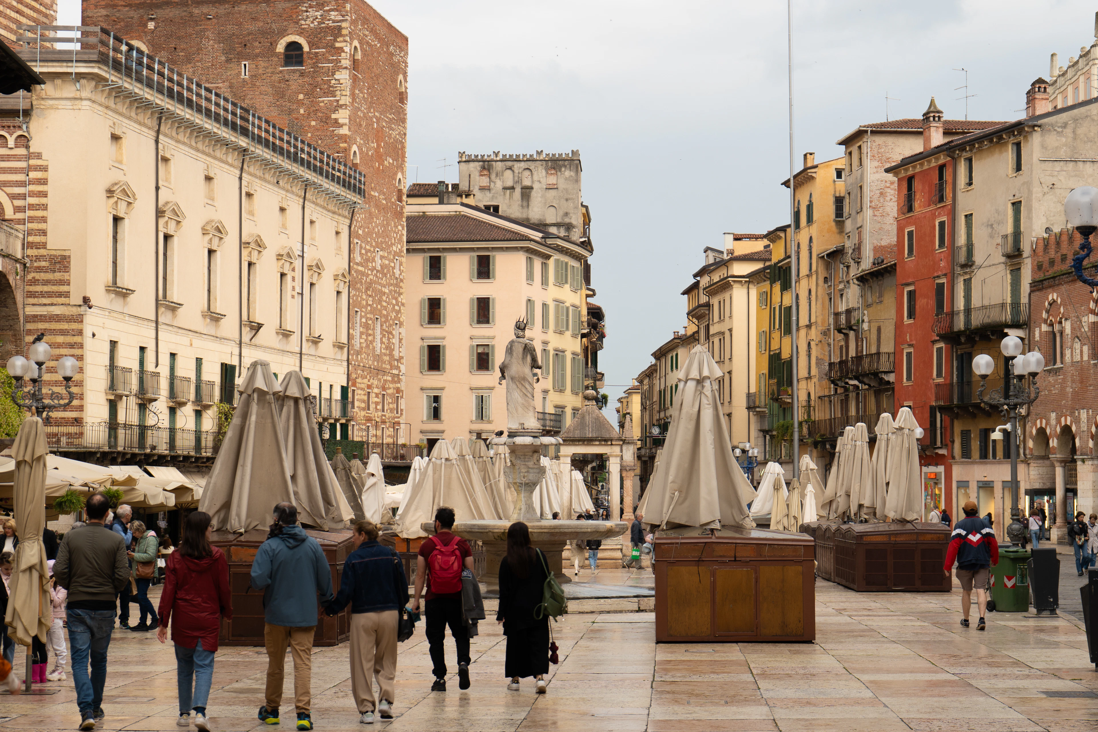 Families walking across Piazza delle Erbe in Verona past the Madonna Verona fountain, with furled market umbrellas, the frescoed facades of the square and a medieval tower closing the view.