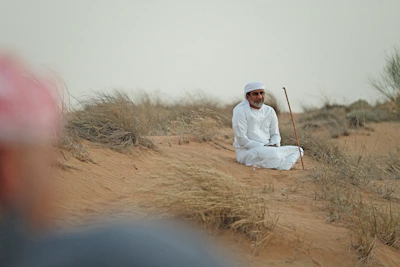 A man sits on the sand in traditional attire.