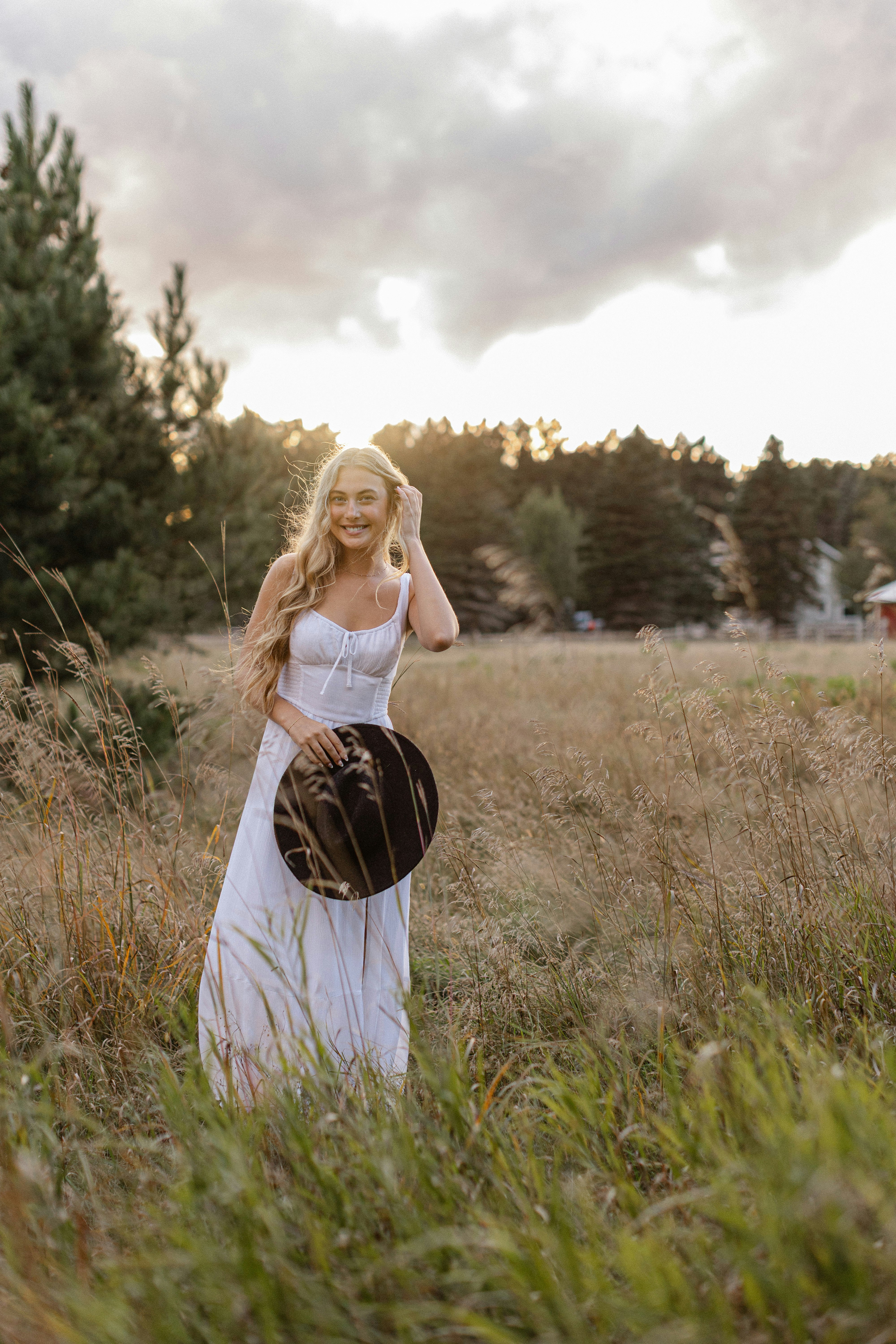 A woman smiles in a field holding a hat.