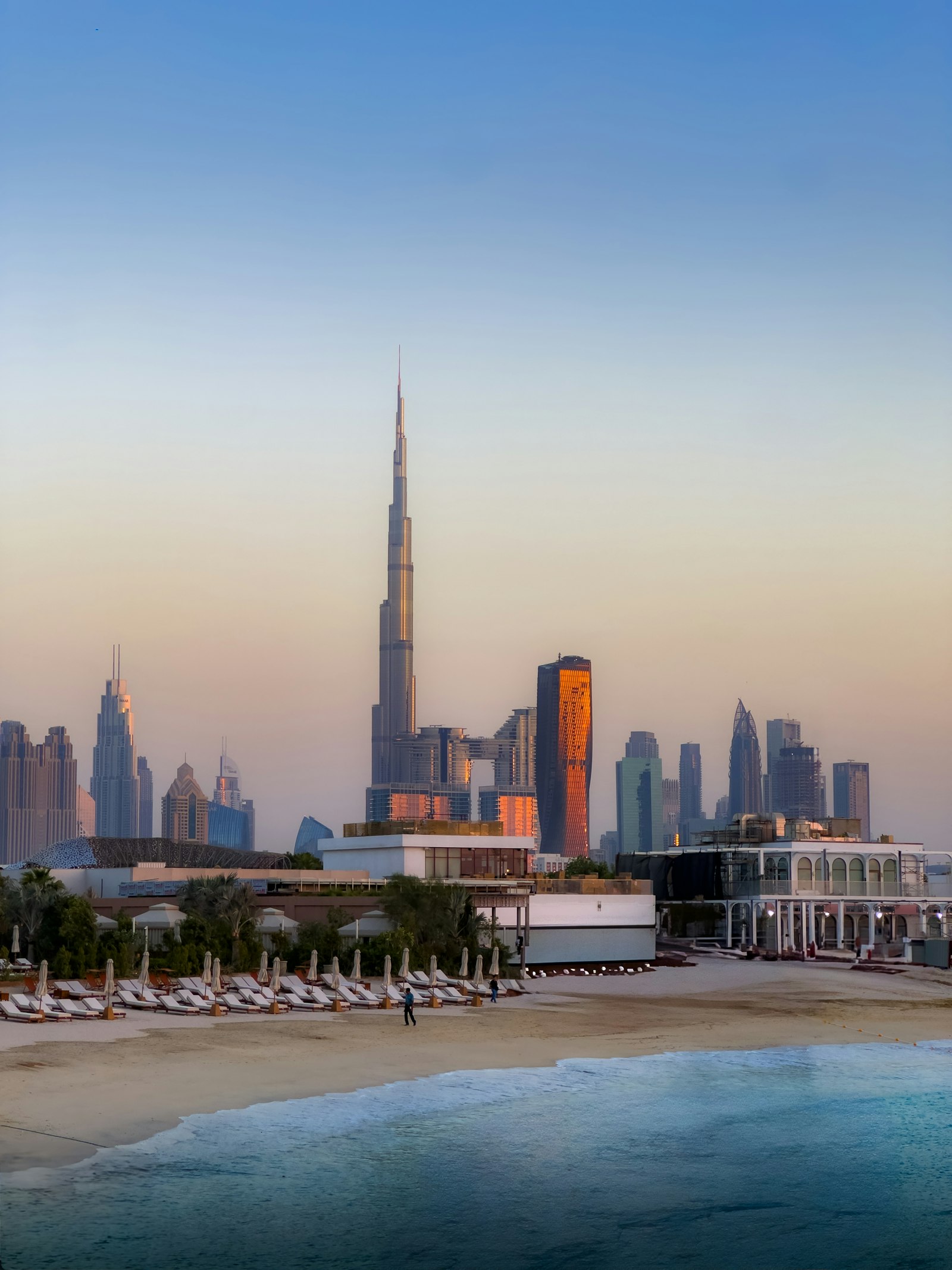 The Dubai skyline and Burj Khalifa seen across the water from a beach at dusk