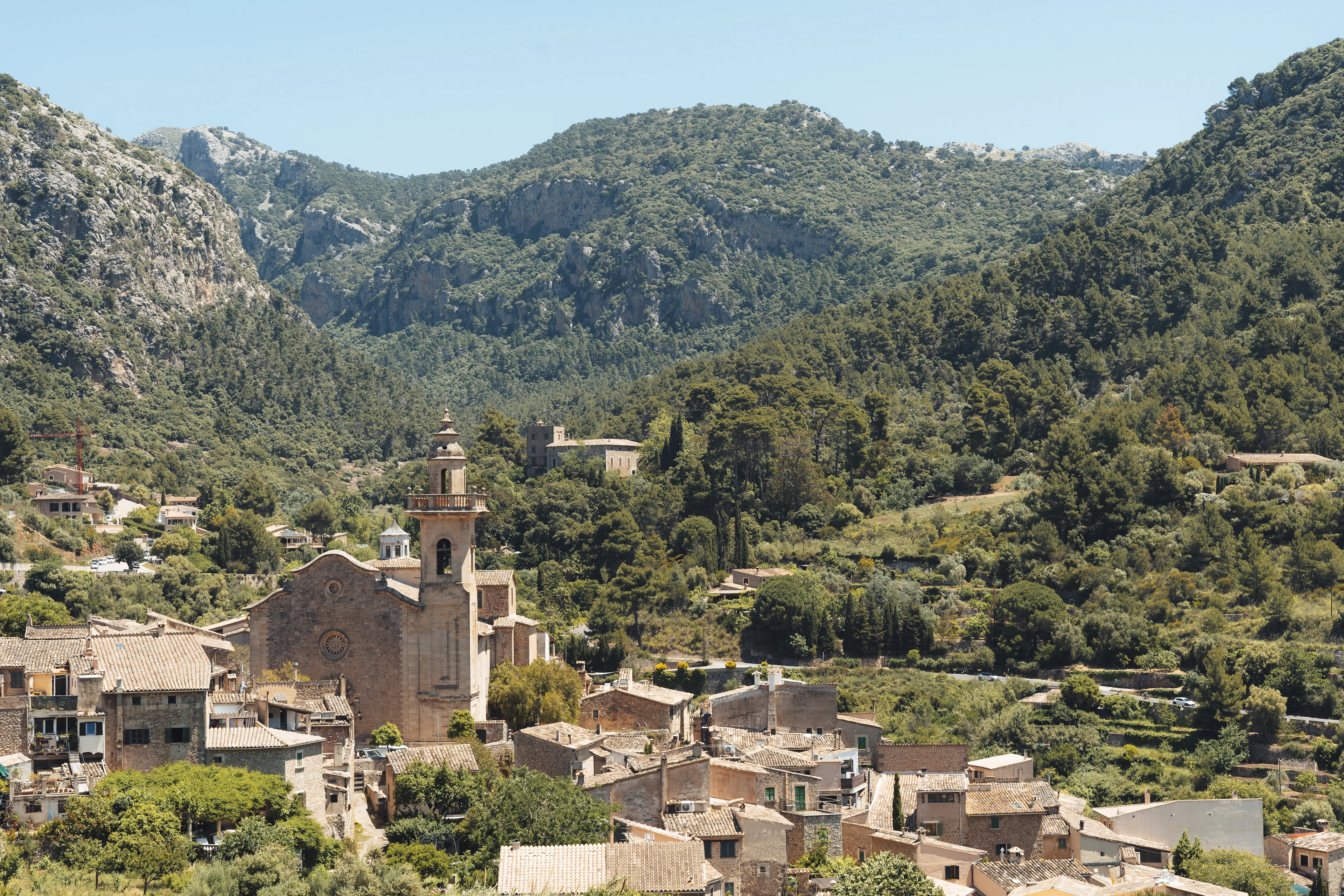 The stone village of Valldemossa on Mallorca, its church tower rising among ochre rooftops on a wooded shoulder of the Serra de Tramuntana under a pale summer sky