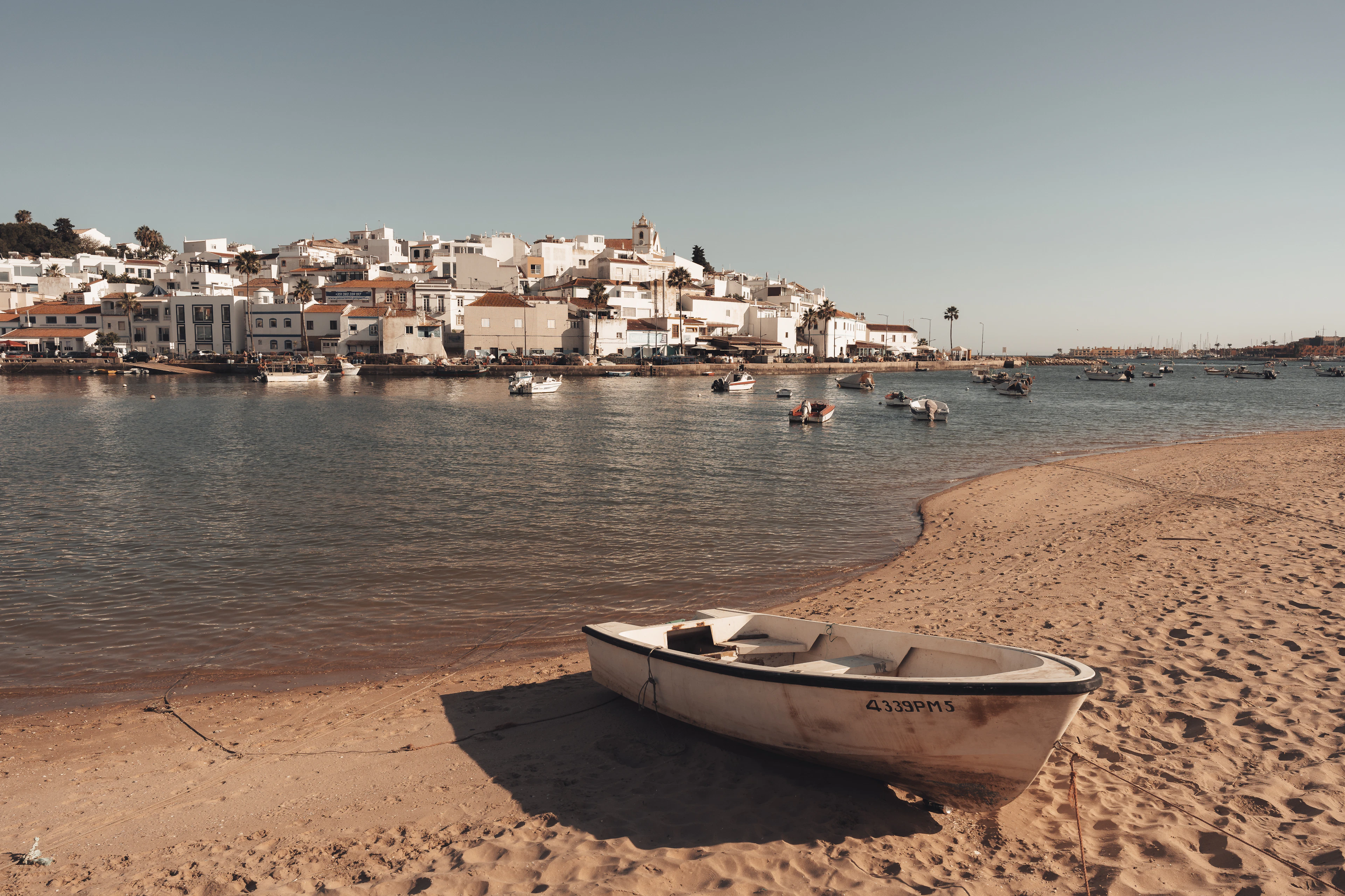 A wooden dinghy resting on the sand at low tide in front of Ferragudo, the whitewashed village stacked up its hillside across the still estuary in warm evening light.