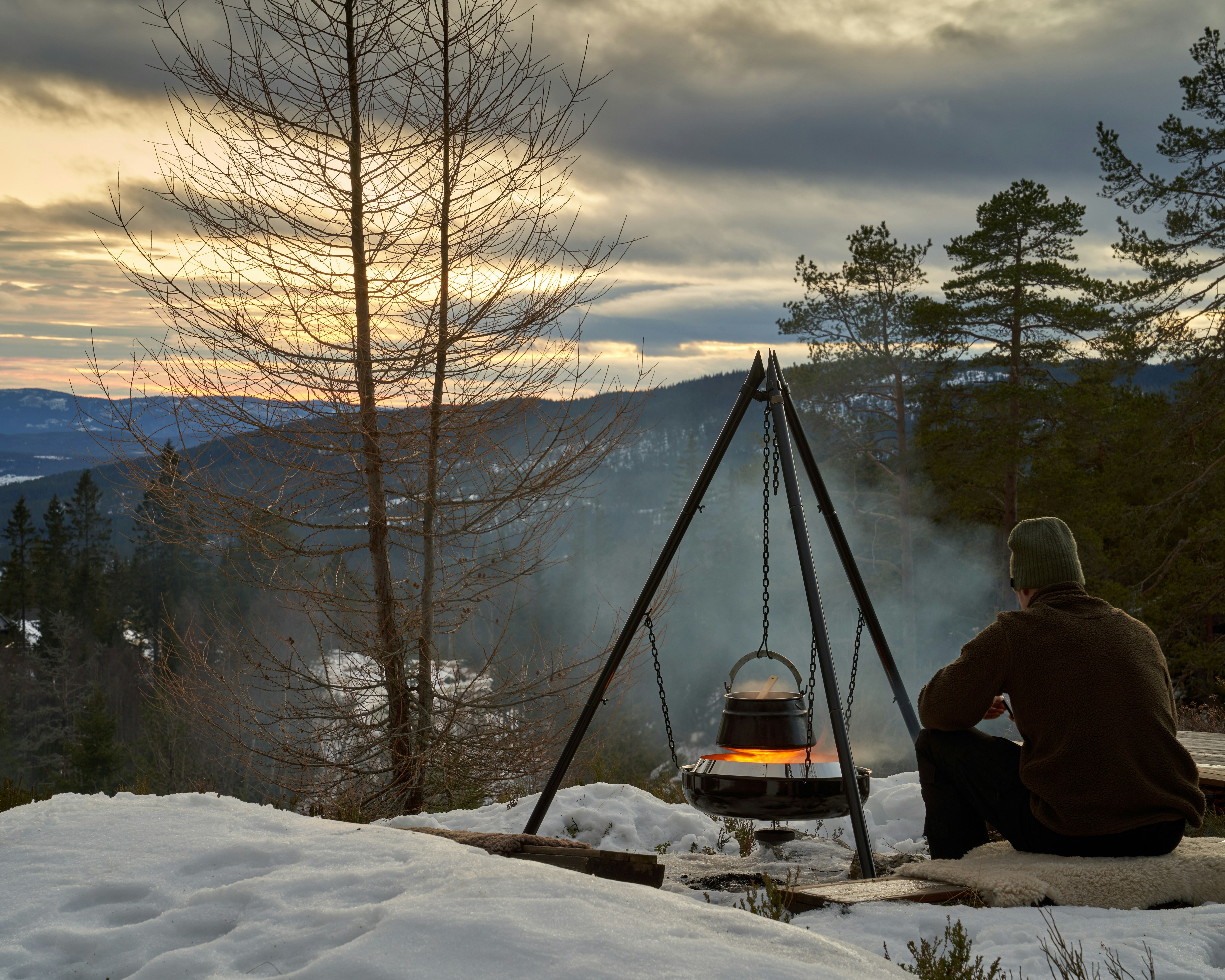 Hungarian goulash is cooking on open fire in the Norwegian woods during winter. View to lake Krøderen during evening light with colorful sunset.