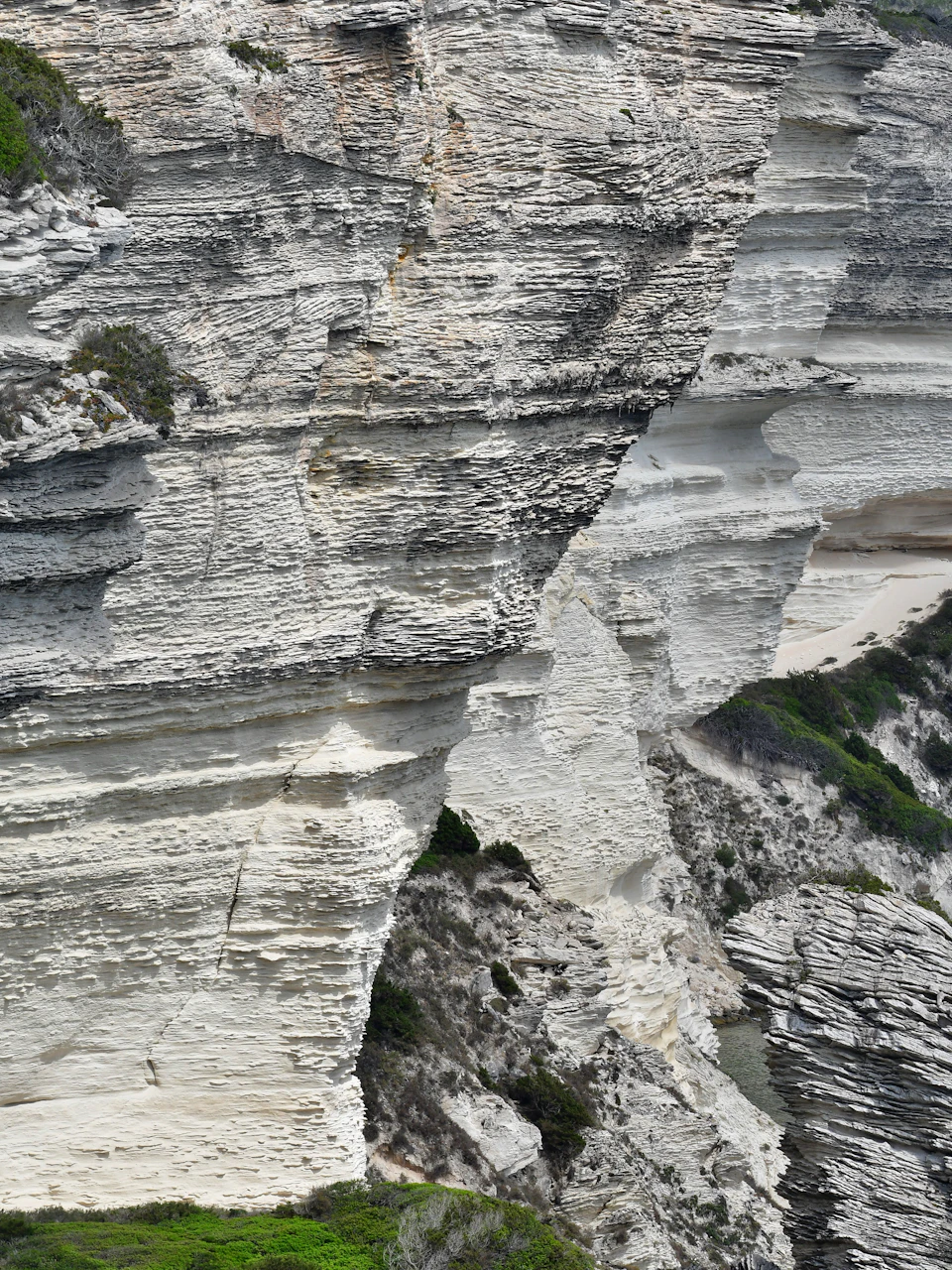 The image shows a close-up view of a cliff face, with white and gray rock layers stacked on top of each other. The rock layers vary in size and shape, creating a sense of depth and texture. In the foreground, there are patches of green vegetation that add a touch of color to the otherwise monochromatic scene.

The image is taken from an elevated perspective, providing a bird's eye view of the cliff face. The background appears to be a light brown color, possibly due to the presence of soil or other natural elements on the cliff face.