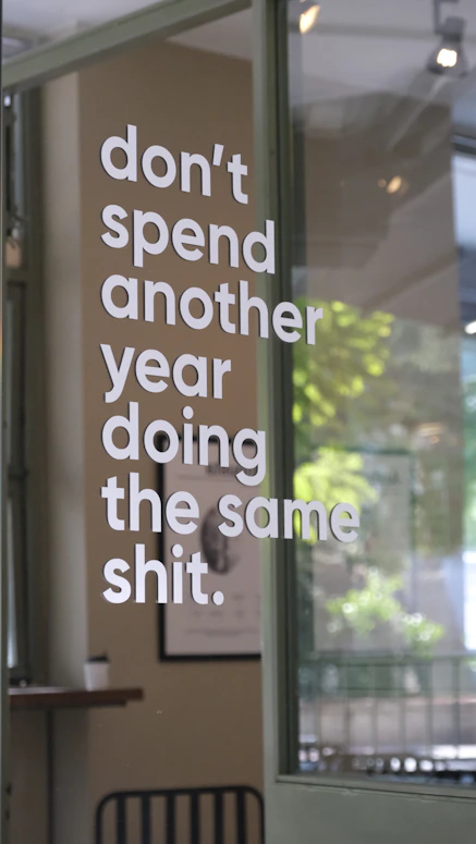 White lettering reading "don't spend another year doing the same shit" on a glass door