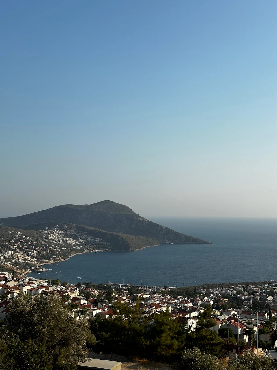 The image presents a panoramic view of a coastal town nestled at the foot of a mountain range, with the ocean stretching out to meet the sky on the horizon. The town is composed of white buildings that blend harmoniously with the surrounding landscape, creating a picturesque scene. The perspective from which the photo is taken allows for an expansive view of the town and its natural surroundings.