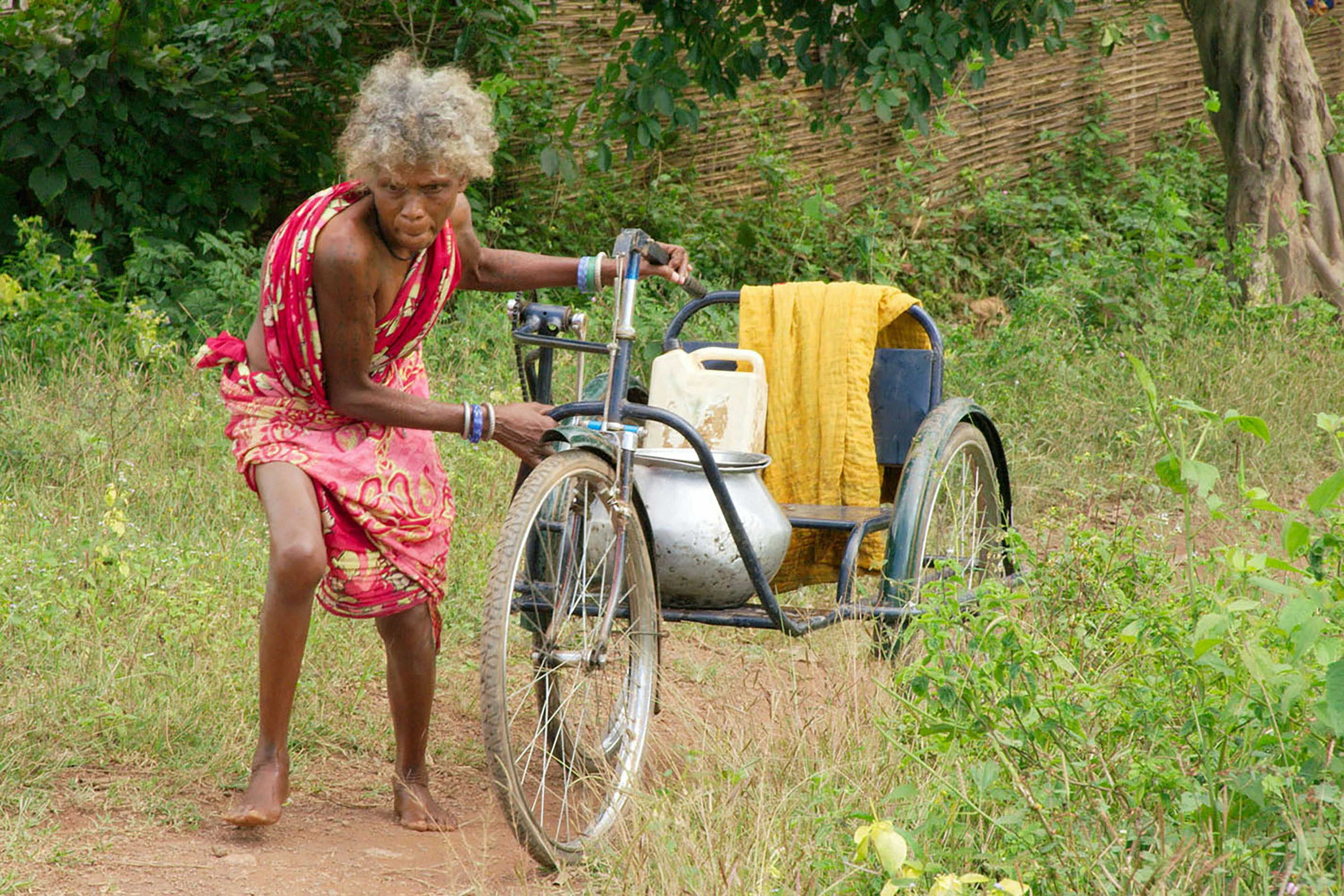 An elderly woman pulls a three-wheeled vehicle.