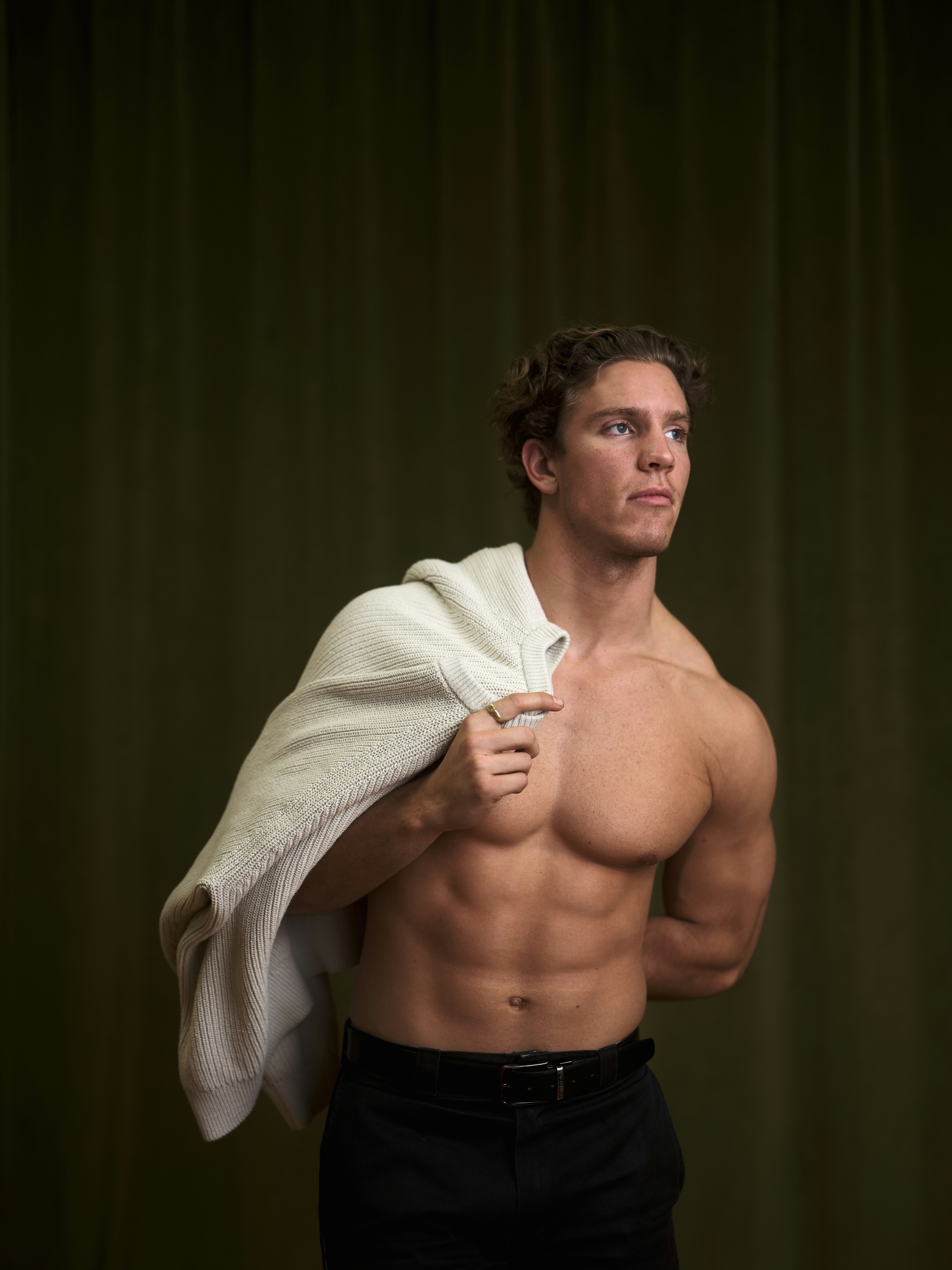 A shirtless man holds a sweater on his shoulder.