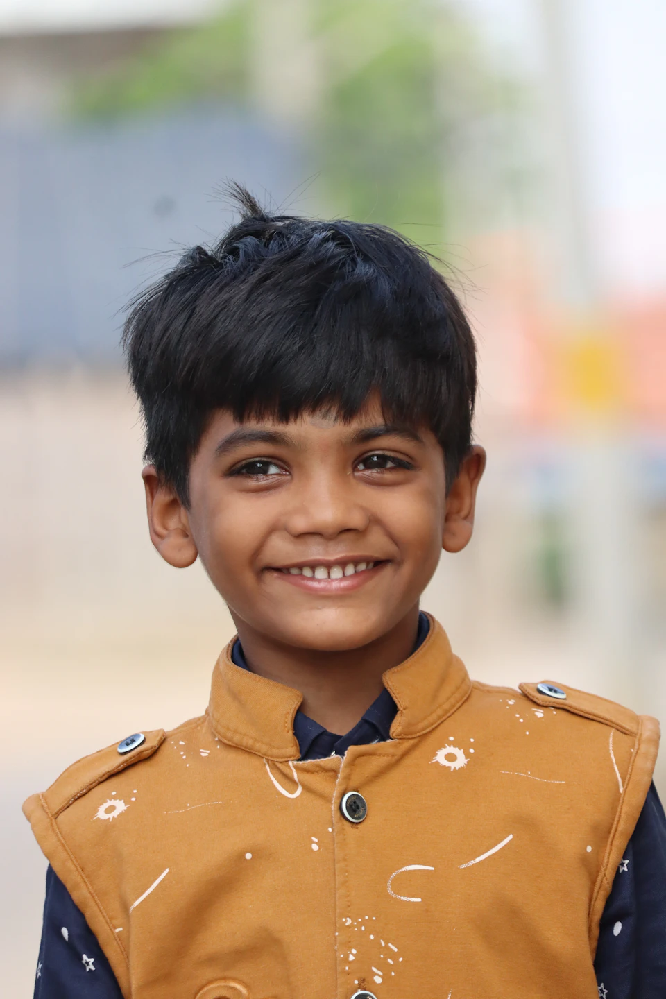 The image shows a young boy with short black hair wearing a brown jacket adorned with white dots and buttons, standing against a blurred background that appears to be an outdoor setting. The boy is looking directly at the camera with a smile on his face, creating a warm and friendly atmosphere in the photo.