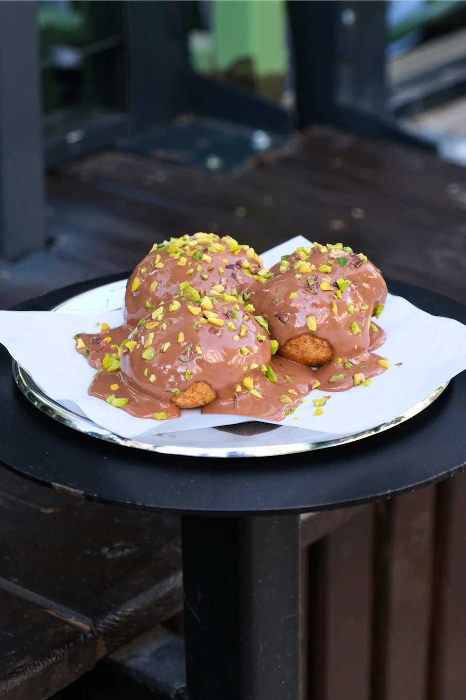 The image shows a plate with three chocolate-covered pastries on a wooden table. The pastries are covered in a layer of chocolate and sprinkled with green pistachio nuts, creating an appetizing visual appeal. The plate is placed on the right side of the frame, drawing attention to its delicious contents.