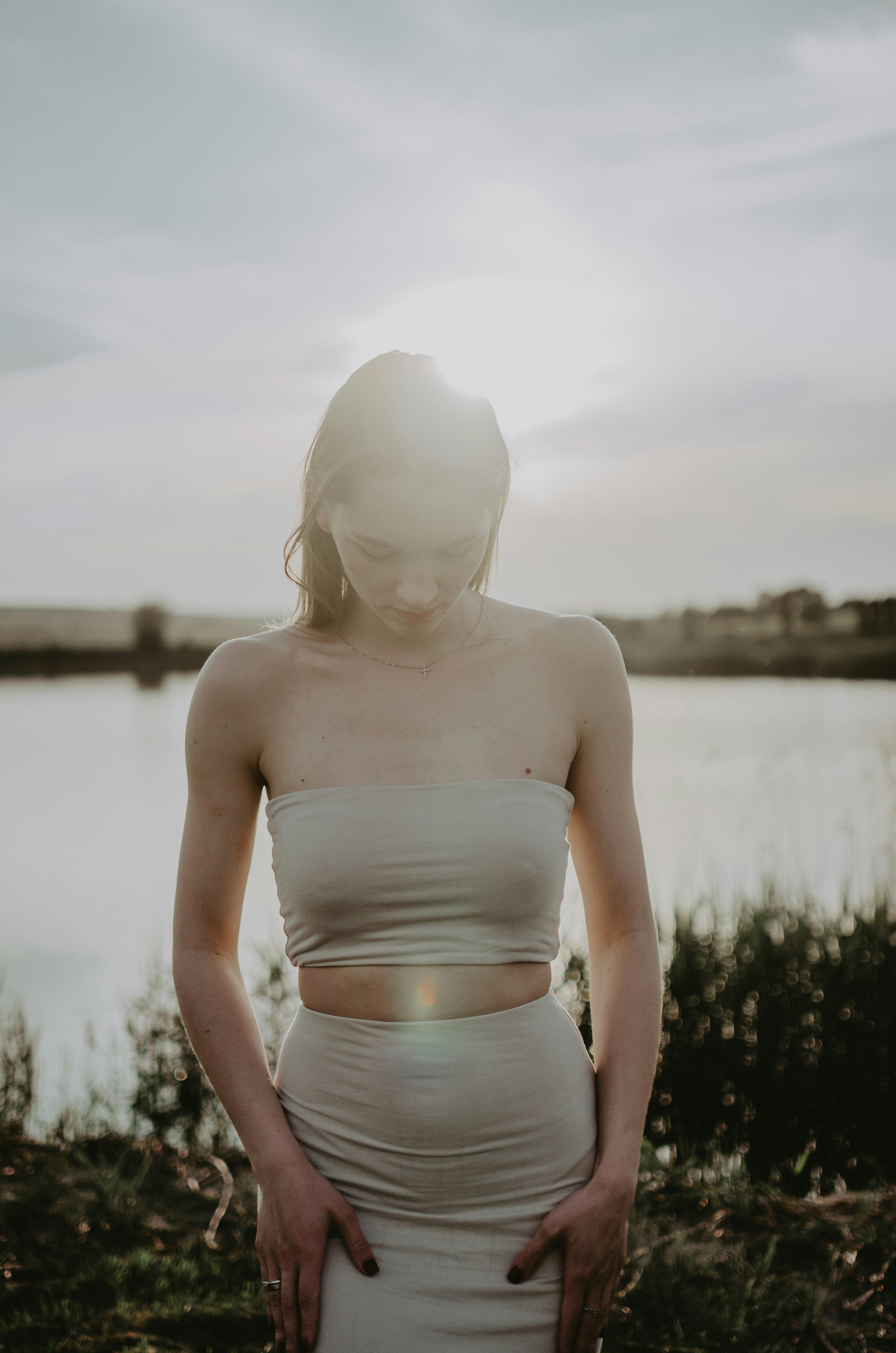 Woman in beige dress looks down by the lake.