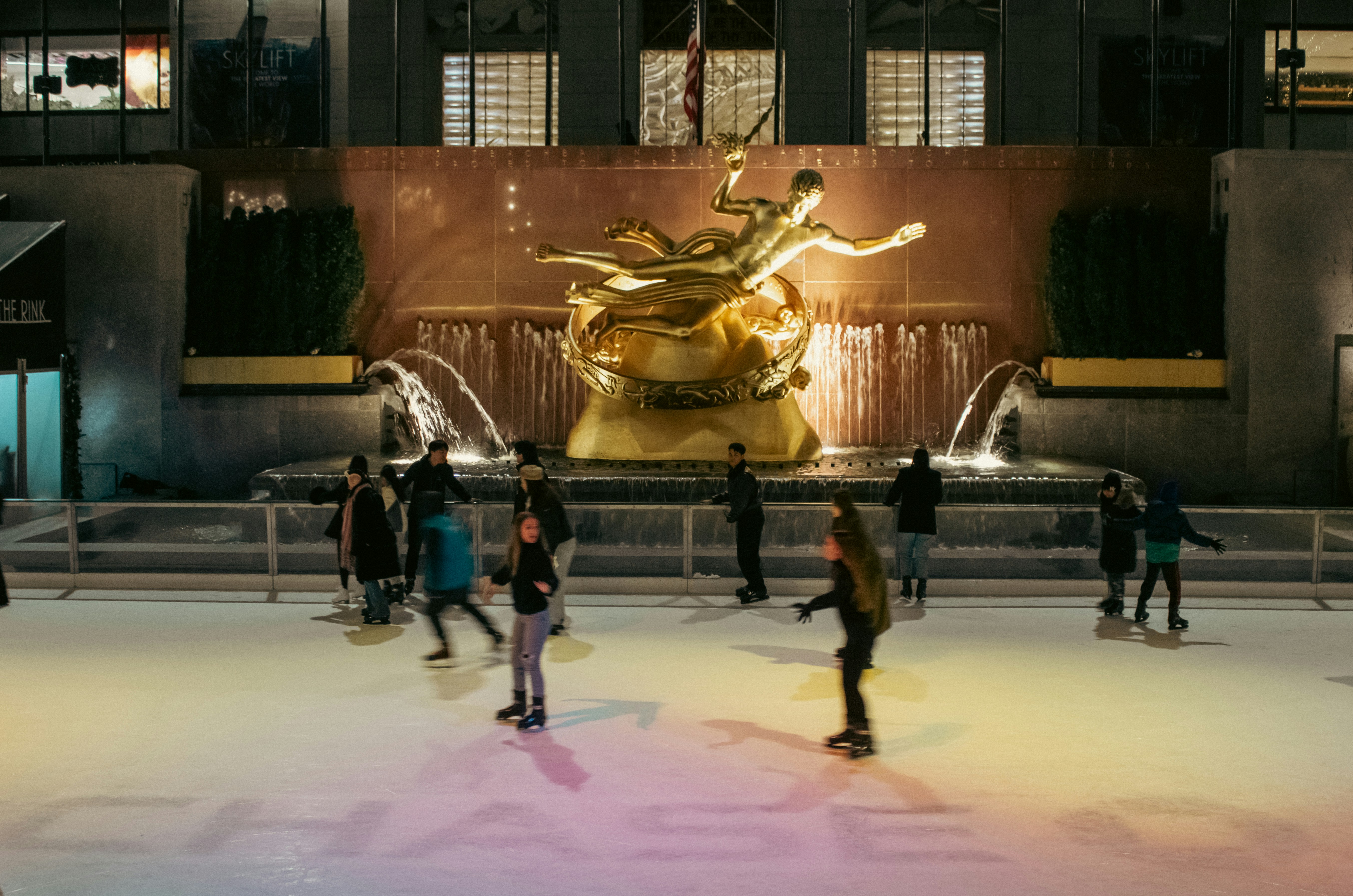 People ice skating at the Rockefeller Center Ice Rink in Midtown Manhattan, New York City. Overlooking the rink is the iconic gilded bronze Prometheus statue by Paul Manship, symbolizing the Titan bringing fire to humanity. Set beneath towering buildings and surrounded by flags and seasonal decor, the rink is a classic winter destination that captures the festive spirit and tradition of NYC.