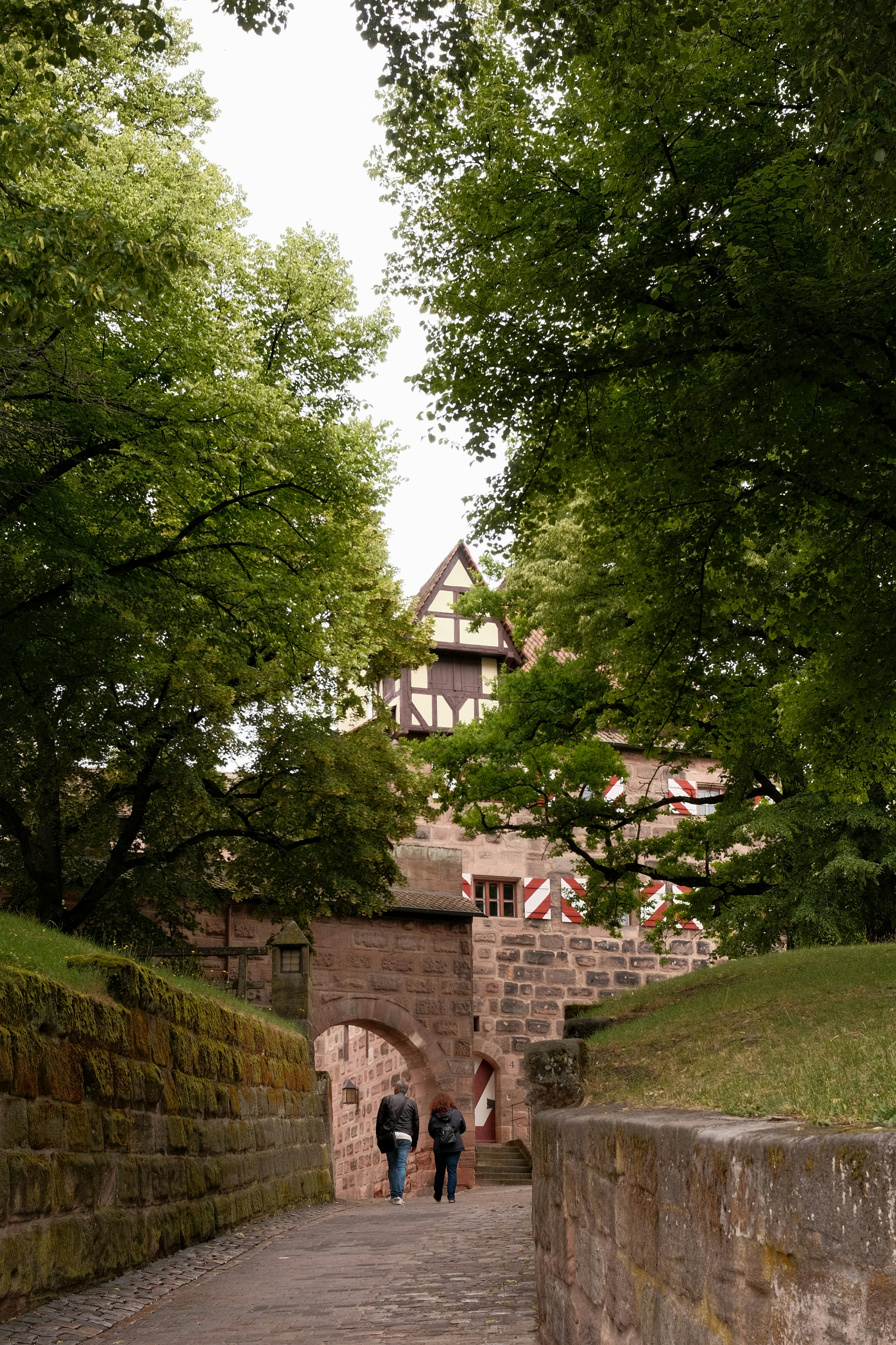 Visitors explore the historic castle grounds surrounded by lush green trees in Nuremberg, Germany, on a cloudy day
