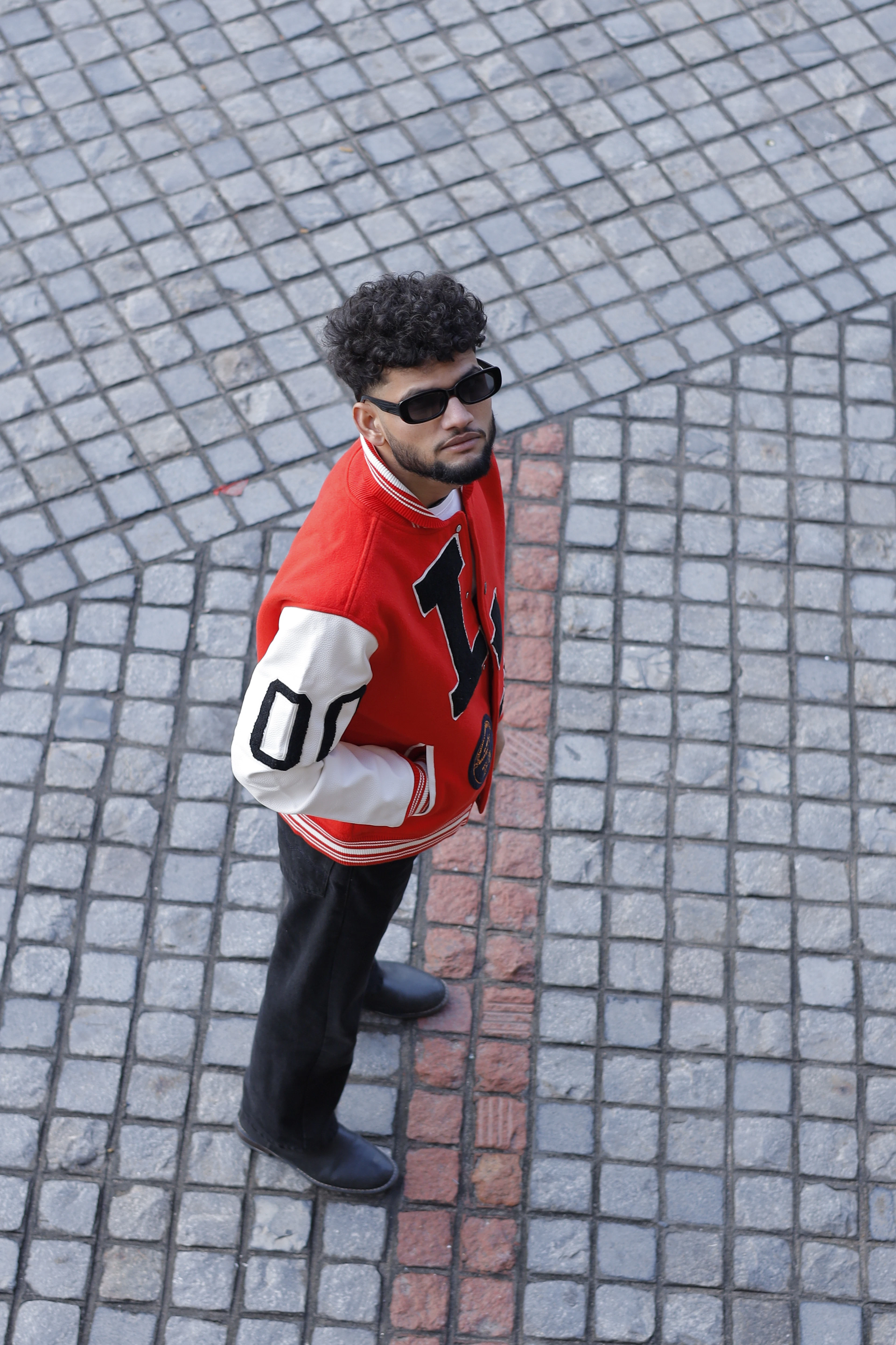 A man in a red and white varsity jacket standing on grey cobblestones, shot from above.