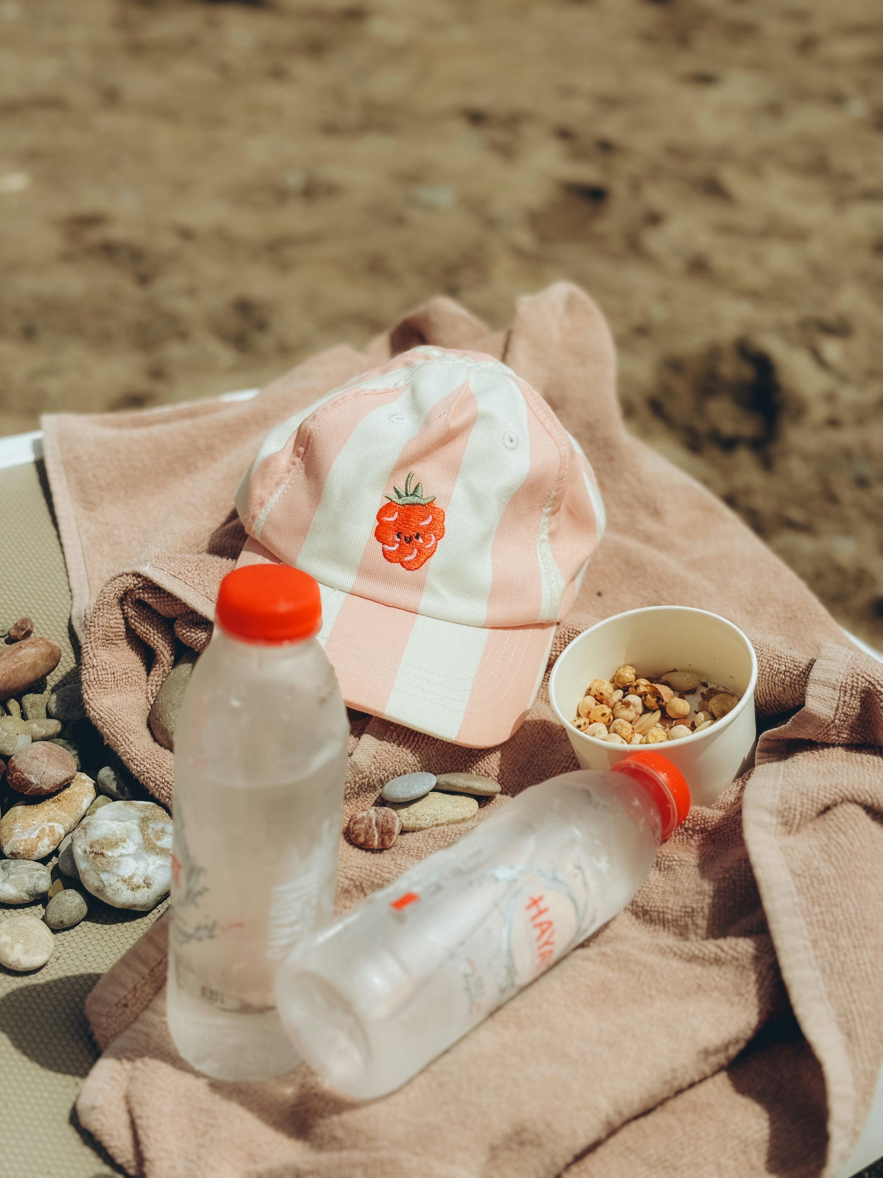 Vivid close-up of a striped pink cap, a small cup of snacks, and two plastic bottles on a towel at the beach, suggesting a casual seaside picnic.